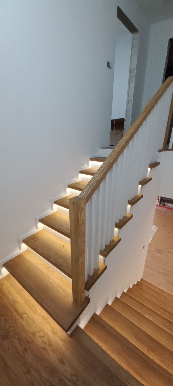Nowoczesne schody z drewna dębowego z podświetleniem LED pod każdym stopniem, biała balustrada z drewnianym poręczą, widok z dołu.