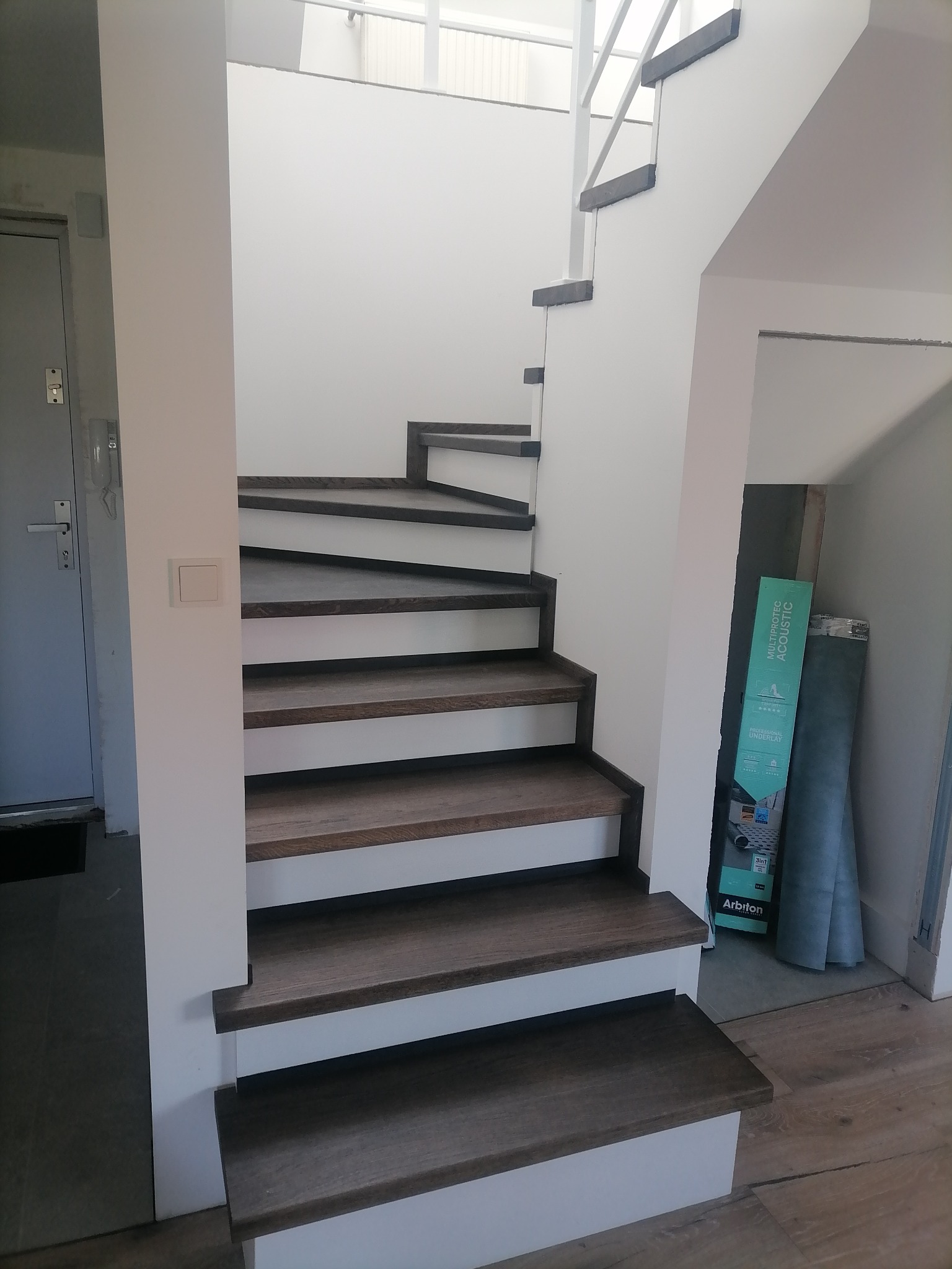 Wewnętrzne schody z drewnianymi stopniami i białymi podstopnicami, widoczne od dołu, z opakowaniem podkładu akustycznego Arbeton w tle.