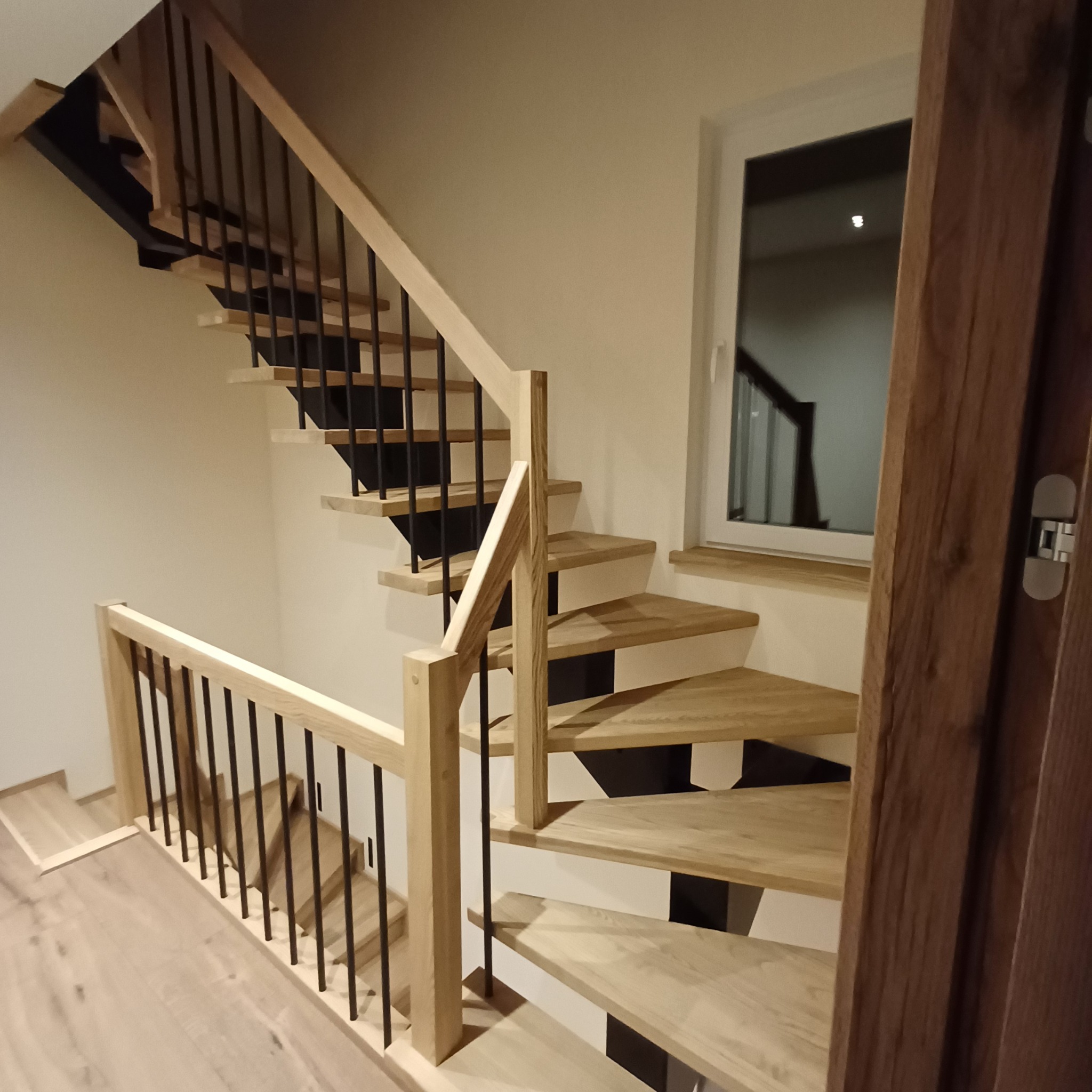 Drewniane schody z jasnego drewna z czarnymi balustradami, widok z dołu, obok okna i fragmentu ciemnych drzwi.