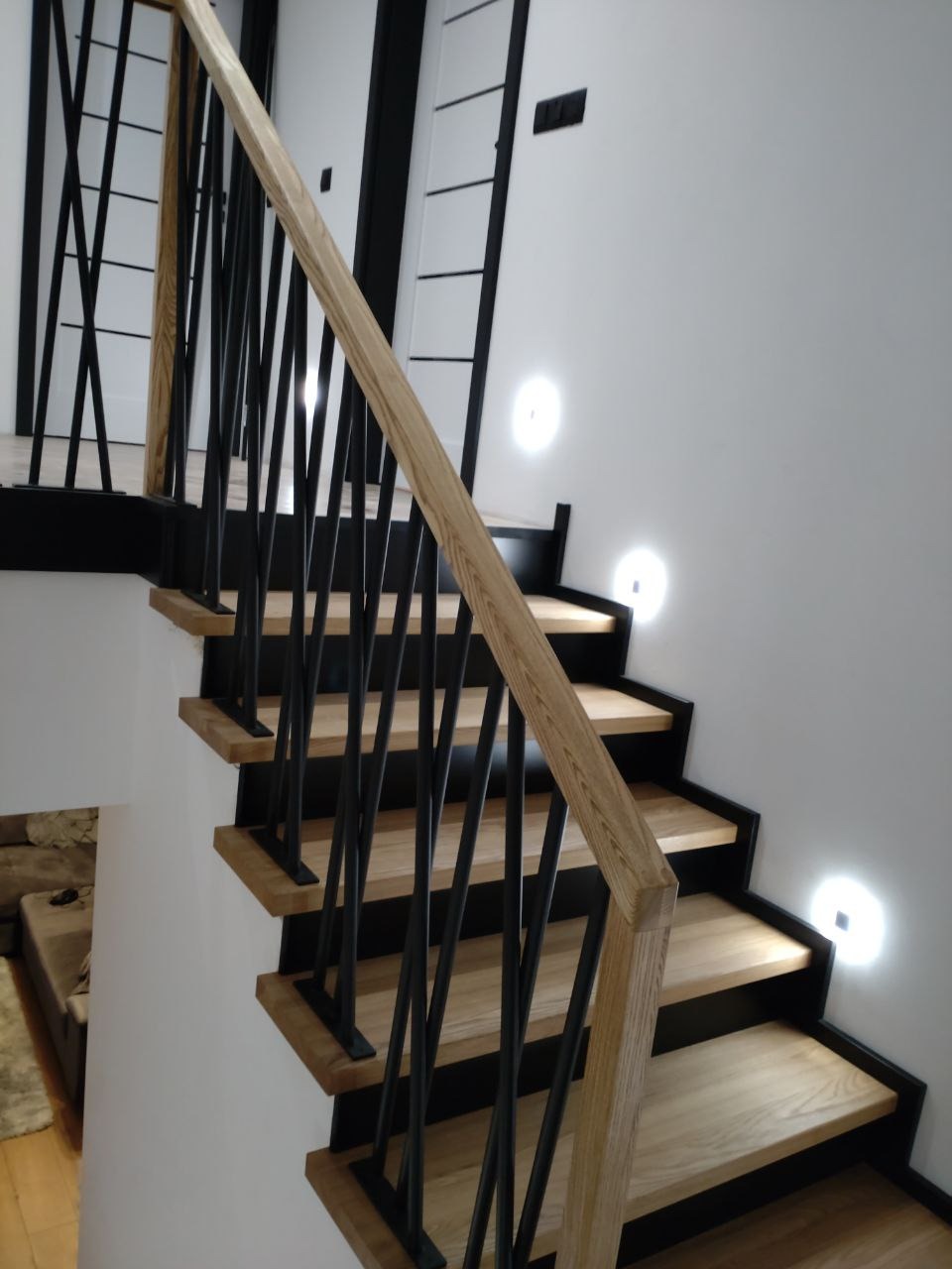 Nowoczesne schody z jasnego drewna z czarnymi elementami konstrukcyjnymi i dyskretnym oświetleniem punktowym na ścianie.