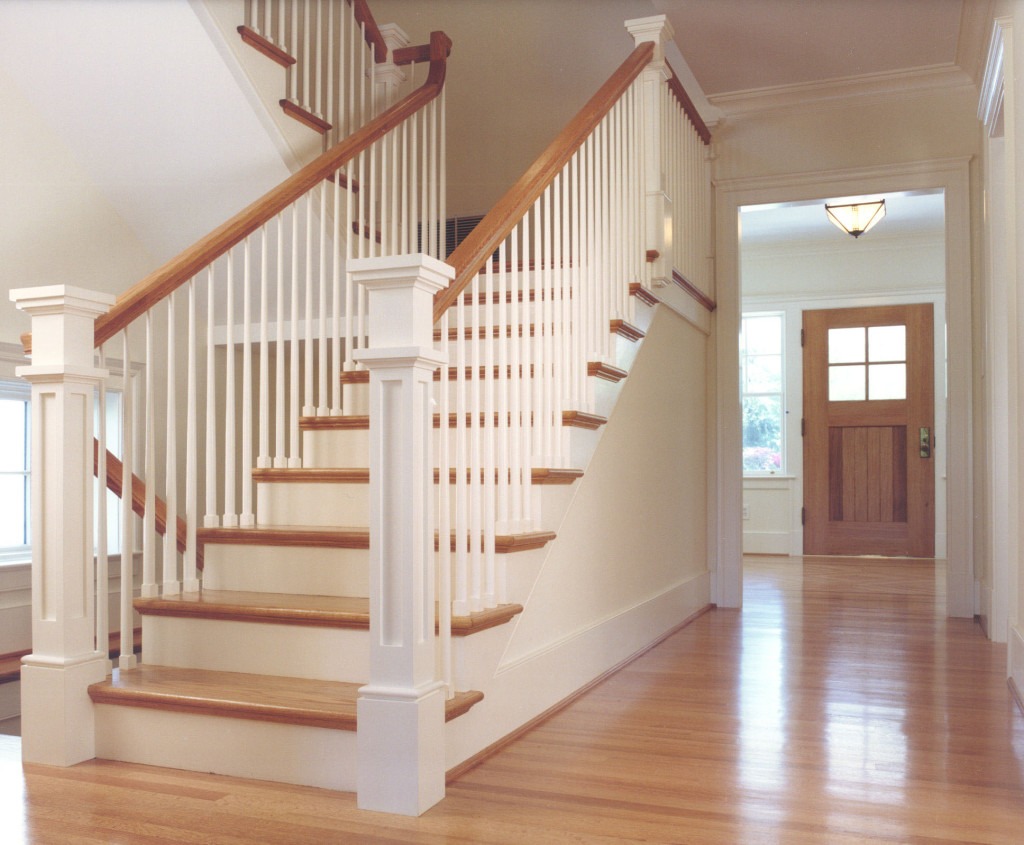 Jasne, klasyczne schody z drewnianymi stopniami i białymi balustradami w przestronnym holu z drewnianą podłogą i widocznymi drzwiami wejściowymi.