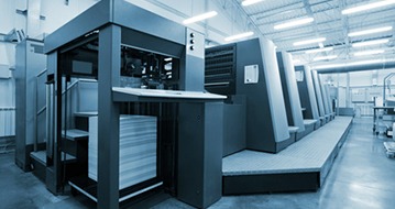 Profesjonalna maszyna drukarska offsetowa w chłodnej tonacji, z widocznym stosem białego papieru i fragmentem hali produkcyjnej w tle.