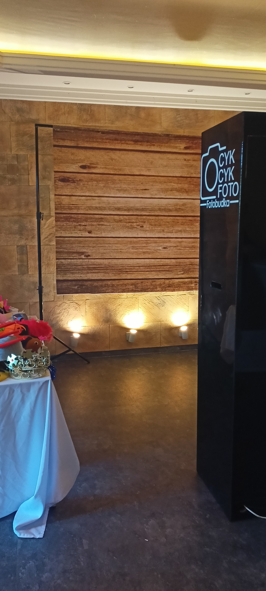 Gotowa do użycia fotobudka z logo 'CYK CYK FOTO' i rekwizytami na stole, ustawiona na tle ściany z drewnianym panelem, oświetlona trzema punktowymi lampami.