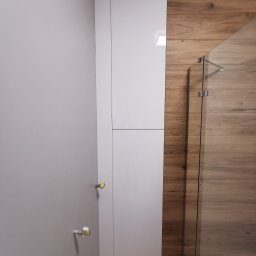 RS Projekt Rafał Szymański - Wysoka, biała szafa na wymiar w łazience z drewnopodobnymi płytkami i szklaną kabiną prysznicową.