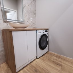 RS Projekt Rafał Szymański - Łazienka z umywalką nablatową w kształcie łodzi, szafką z frontami w kolorze białym i drewnopodobnym blatem oraz pralką LG obok szafki; ściana wyłożona płytkami imitującymi marmur.