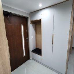 RS Projekt Rafał Szymański - Wnętrze przedpokoju z zabudową: szara szafa z drewnianym akcentem, wbudowane siedzisko z wieszakami i brązowe drzwi wejściowe z przeszkleniem.