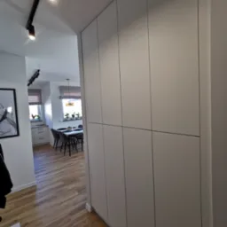 Wysoka, zabudowana szafa wnękowa w przedpokoju, z widokiem na jadalnię. Minimalistyczny design, jasne fronty, oświetlenie szynowe na suficie. Parkiet ułożony w jodełkę.