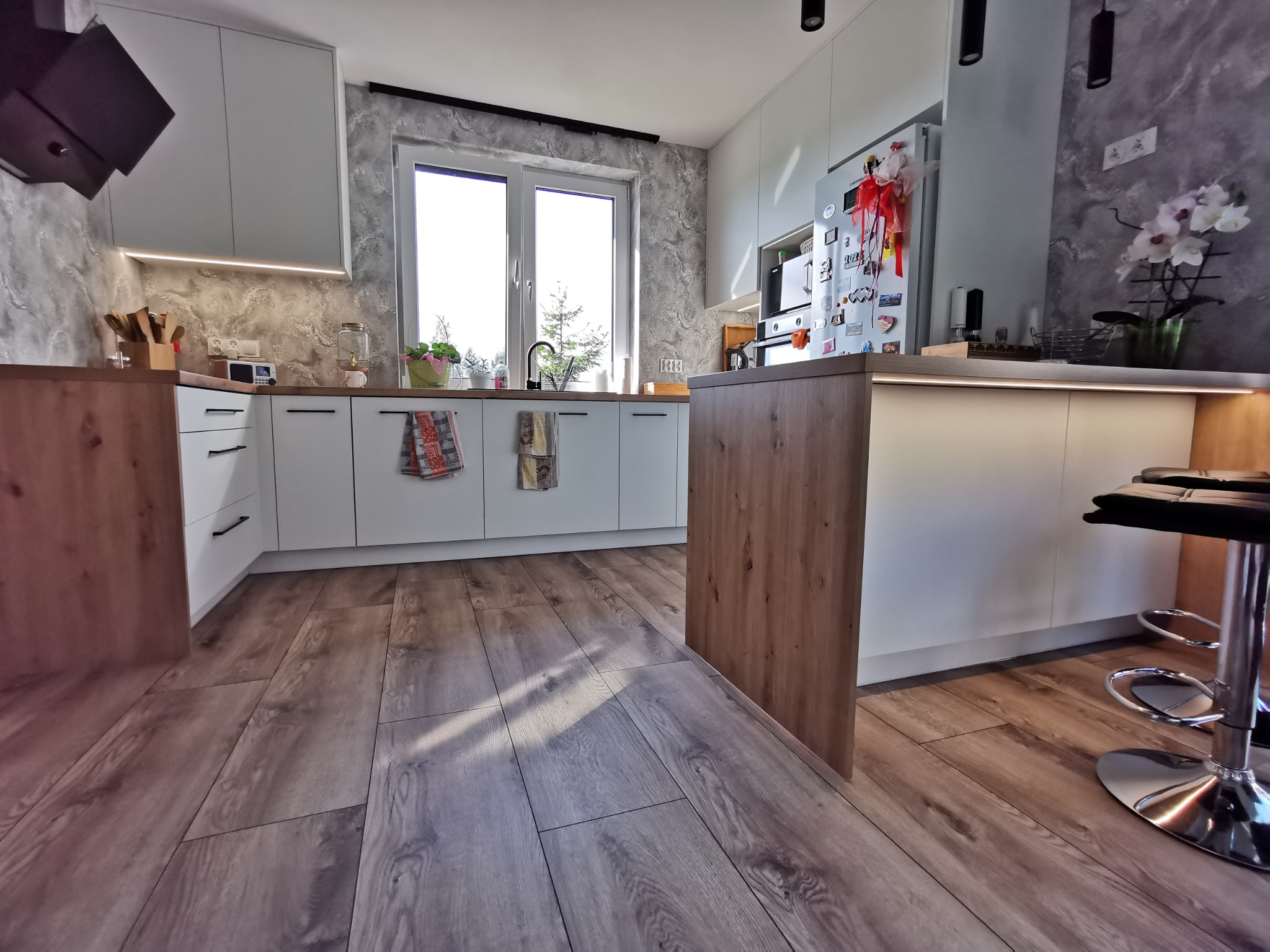 Jasna kuchnia z drewnianym blatem i wyspą, białe fronty szafek, okno z widokiem na zieleń, drewniana podłoga, nowoczesne oświetlenie podszafkowe, krzesło barowe z chromowaną podstawą.