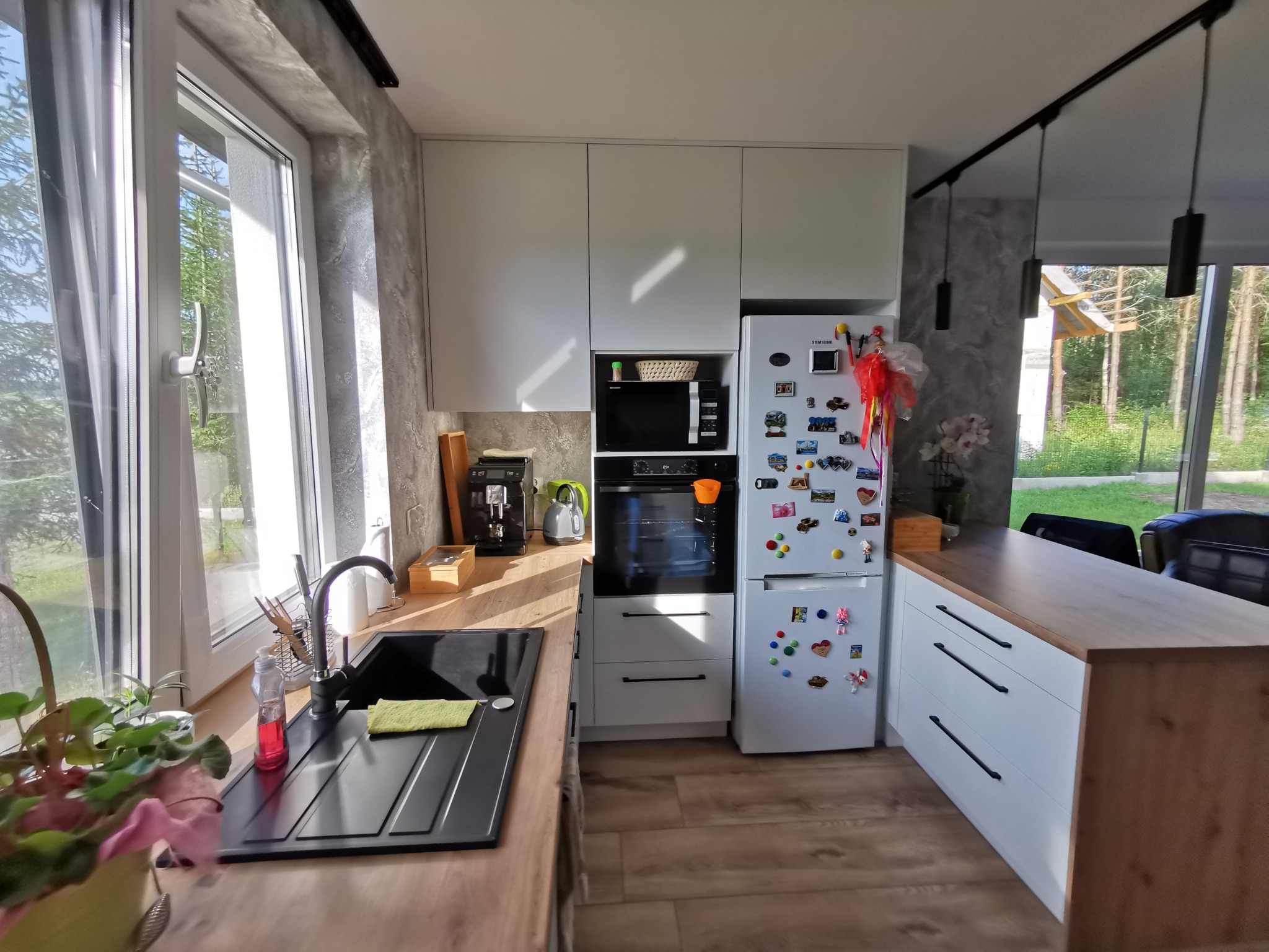 Jasna kuchnia z drewnianym blatem i czarnym zlewem pod oknem, białe szafki wiszące i stojące z wbudowanym piekarnikiem i mikrofalówką, lodówka ozdobiona magnesami, podłoga z paneli imitujących drewno.