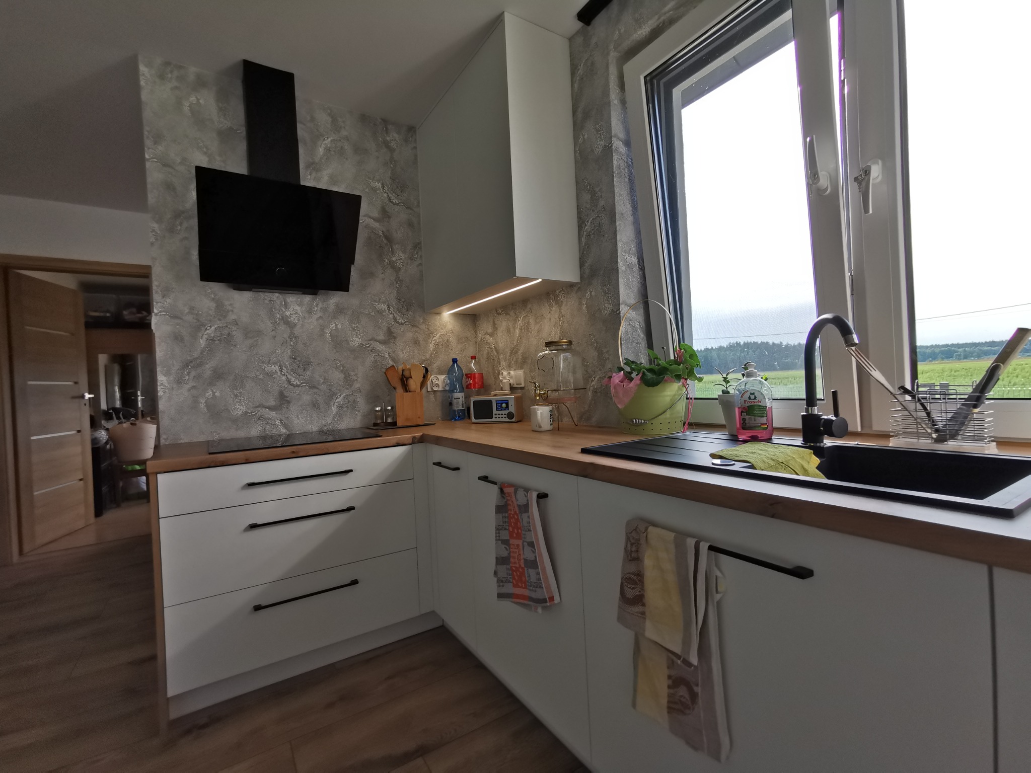 Nowoczesna kuchnia w odcieniach bieli i szarości z drewnianym blatem, czarnym zlewem i armaturą, oraz dekoracyjną ścianą z efektem betonu.