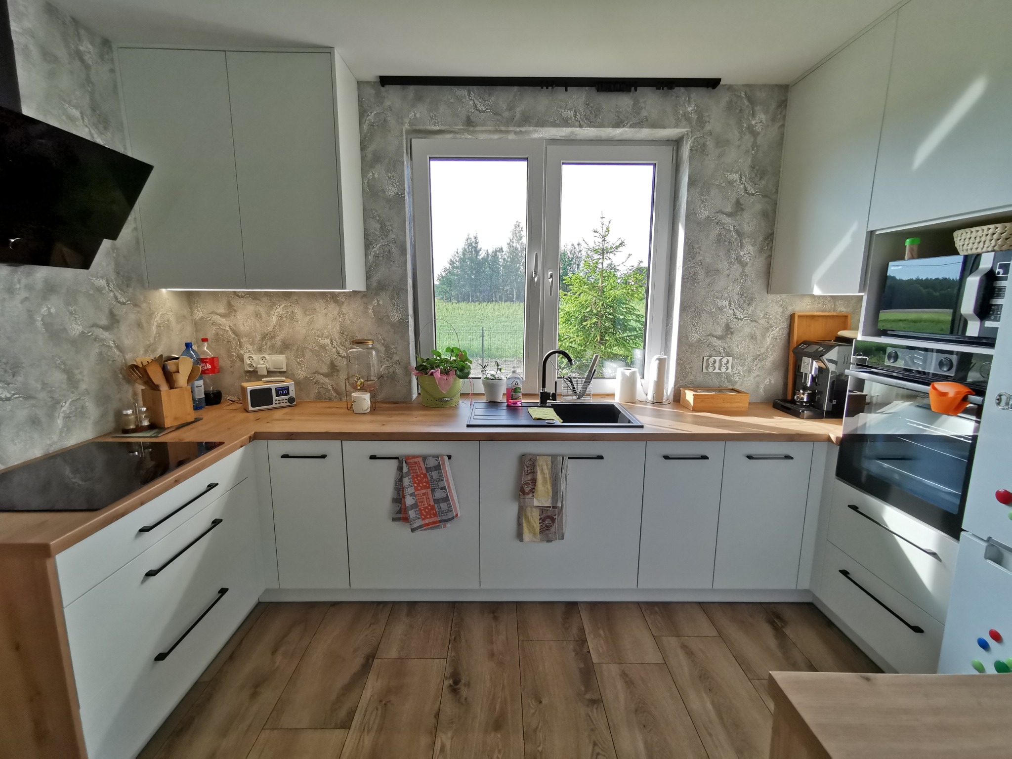 Jasna kuchnia z białymi szafkami i drewnianym blatem, czarny zlew pod oknem z widokiem na zieleń, drewniana podłoga, nowoczesny design z długimi, czarnymi uchwytami.