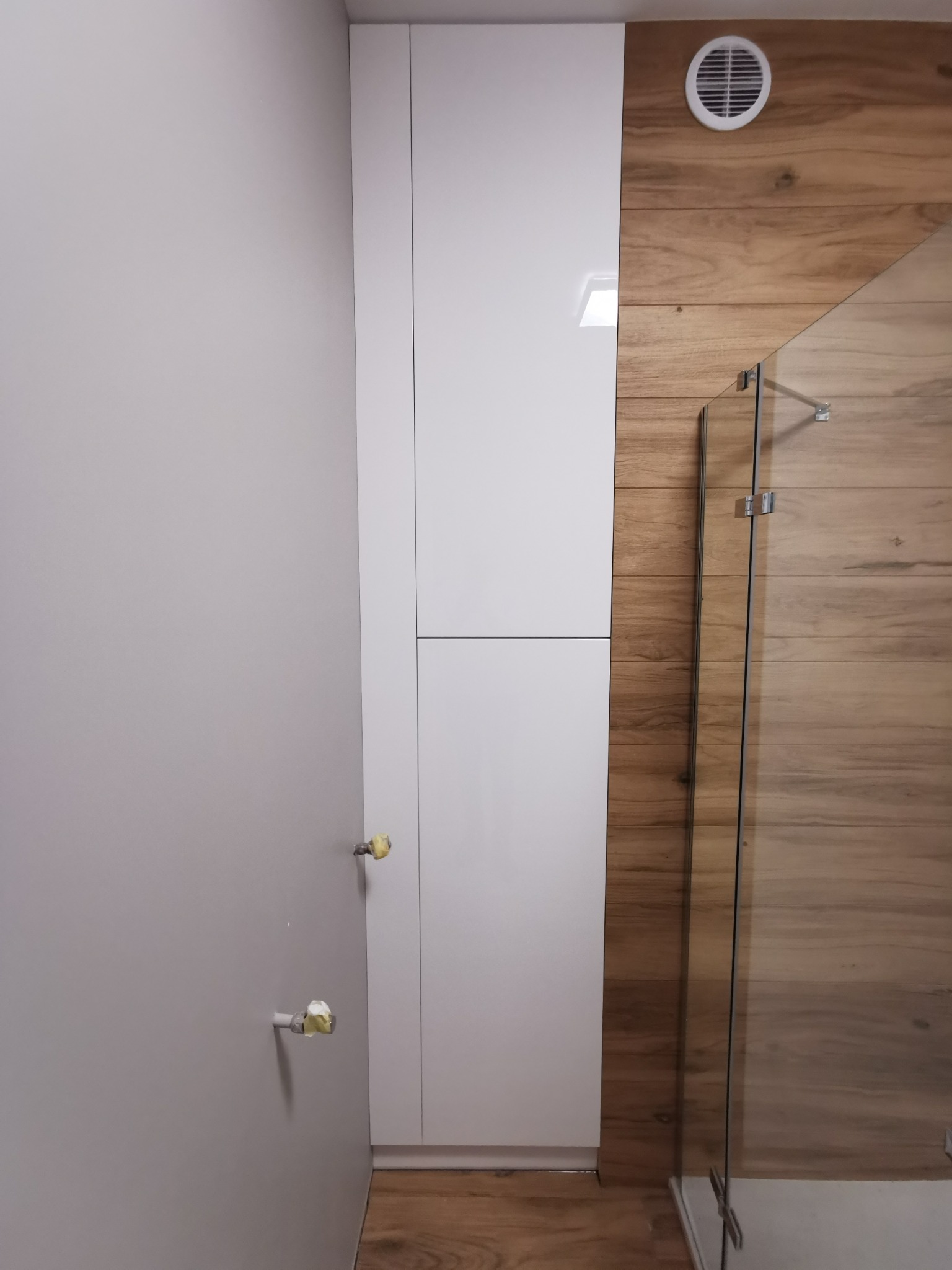 Wysoka, biała szafa na wymiar w łazience z drewnopodobnymi płytkami i szklaną kabiną prysznicową.