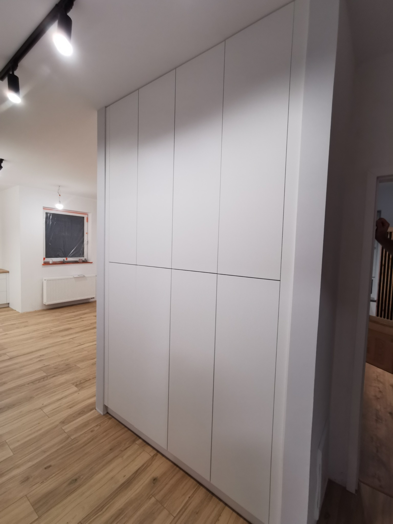 Biała szafa wnękowa z prostymi frontami, zamontowana w korytarzu z drewnopodobną podłogą, widok na remontowane pomieszczenie z oknem zabezpieczonym folią.
