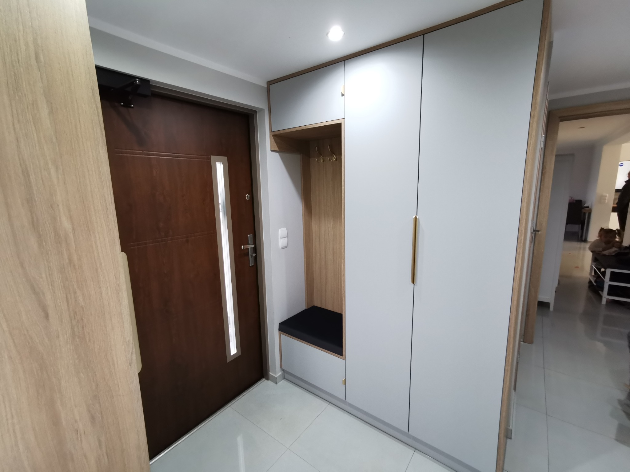 Wnętrze przedpokoju z zabudową: szara szafa z drewnianym akcentem, wbudowane siedzisko z wieszakami i brązowe drzwi wejściowe z przeszkleniem.