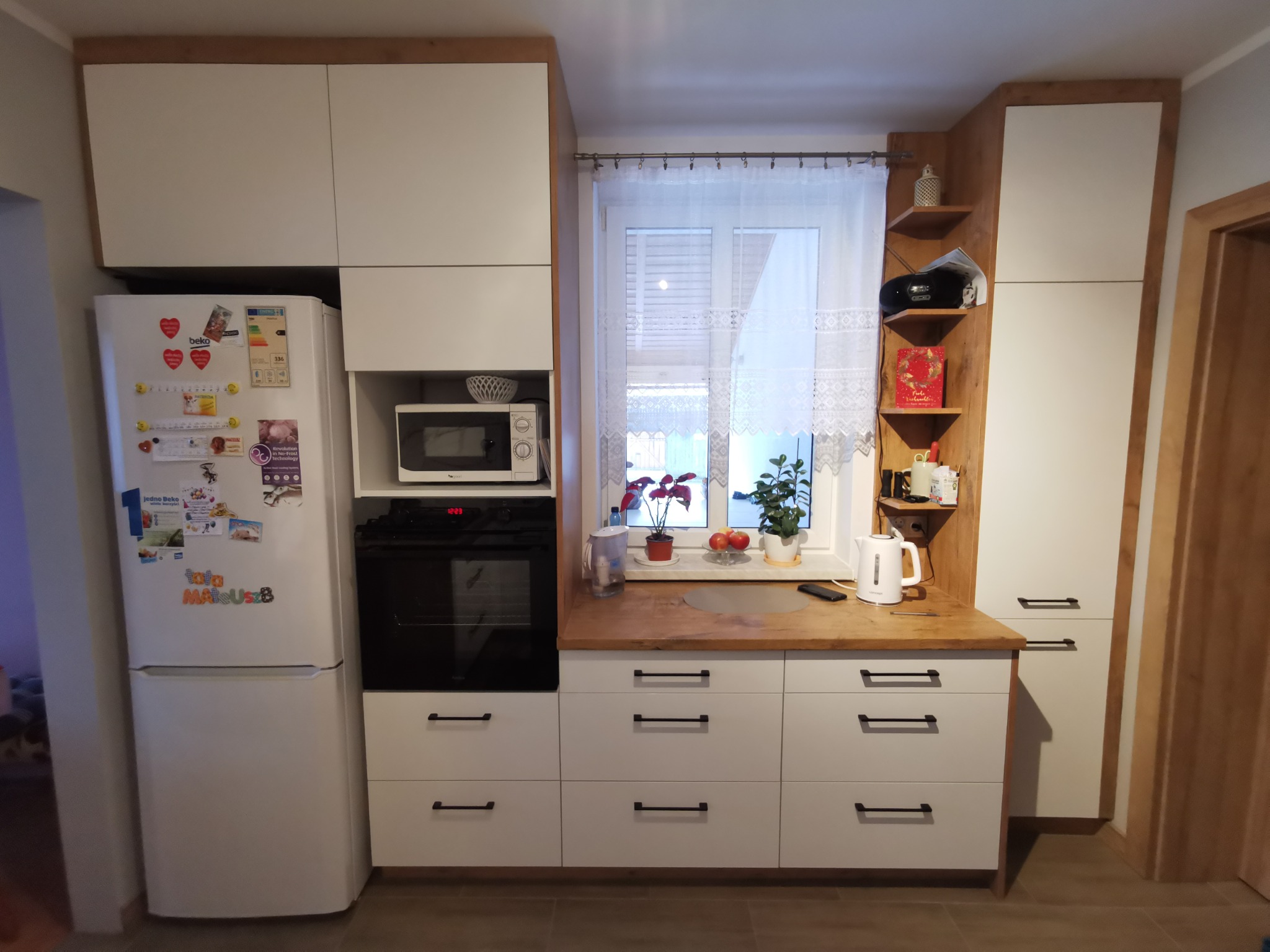 Jasna kuchnia z białymi frontami i drewnianymi akcentami, wyposażona w lodówkę z magnesami, piekarnik, mikrofalówkę i szafki z czarnymi uchwytami, oświetlona naturalnym światłem z okna z firanką.