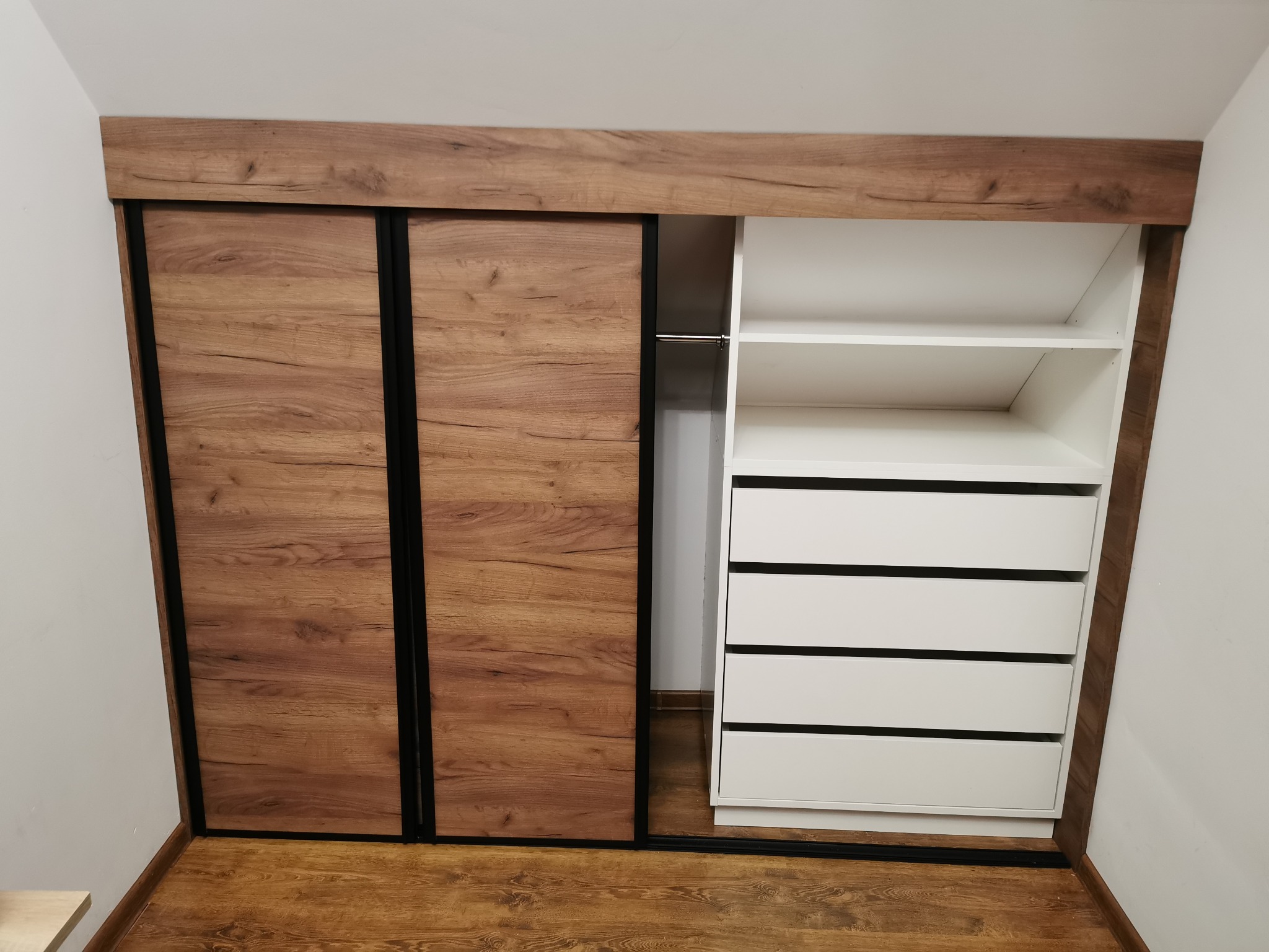 Szafa wnękowa z przesuwnymi drzwiami z okleiną drewnopodobną w kolorze orzecha, z czarnymi obramowaniami, po prawej stronie białe półki i szuflady, dopasowana do skosu poddasza.