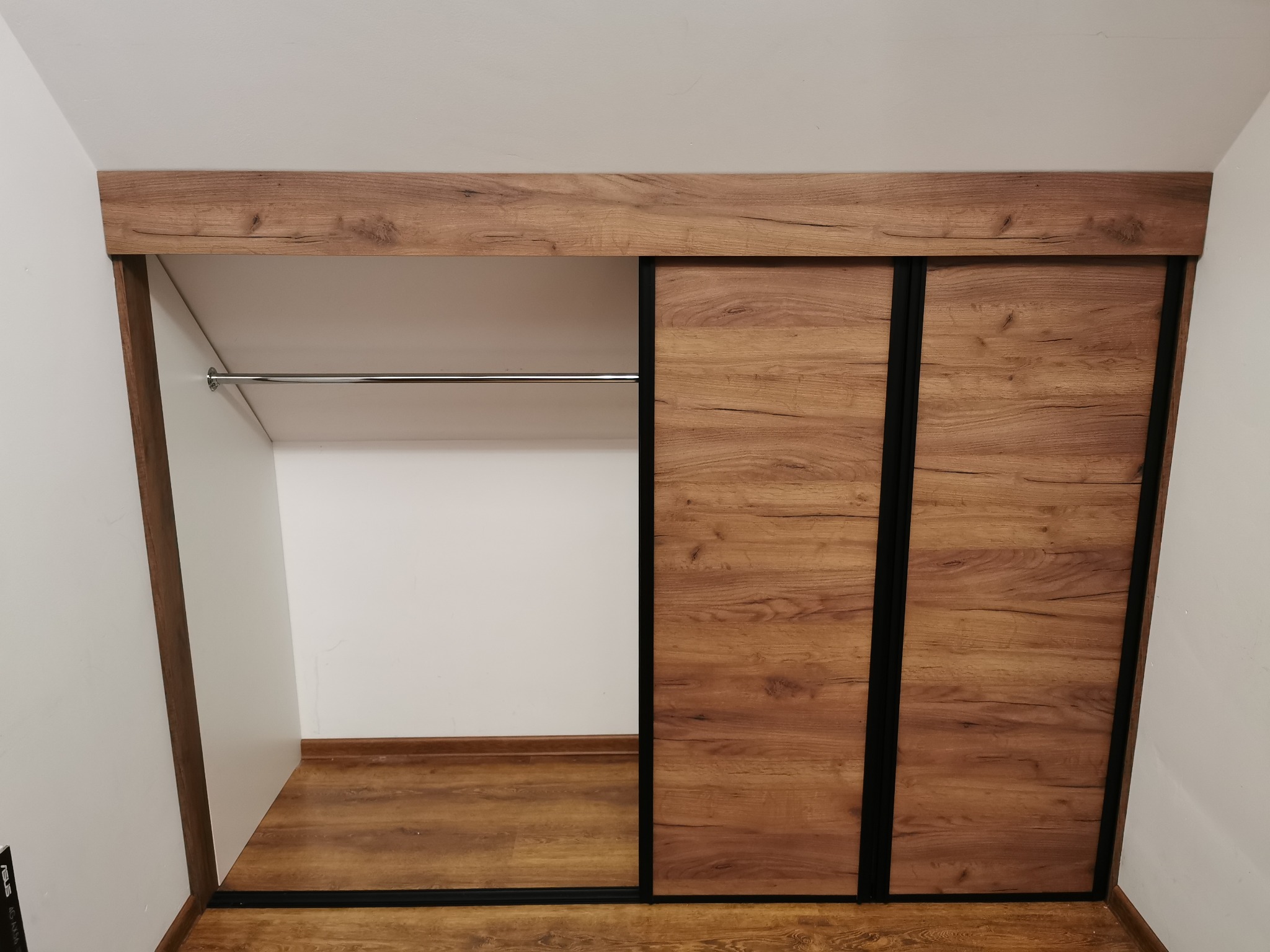 Szafa wnękowa z przesuwnymi drzwiami o fakturze drewna, czarne obramowanie, metalowy drążek na ubrania w otwartej części.