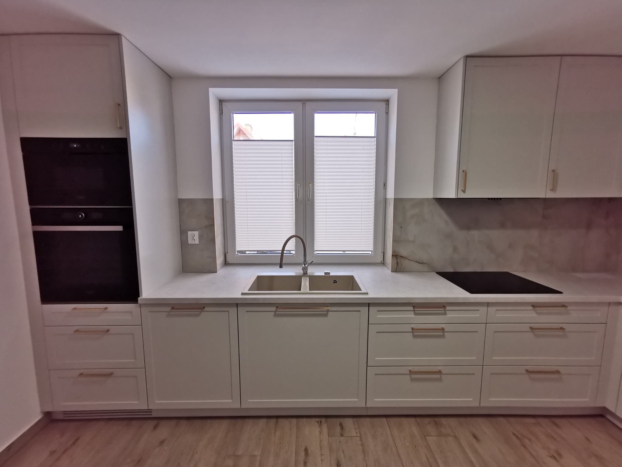 Jasna kuchnia na wymiar z białymi frontami, złotymi uchwytami, blatem imitującym marmur i oknem z plisami.