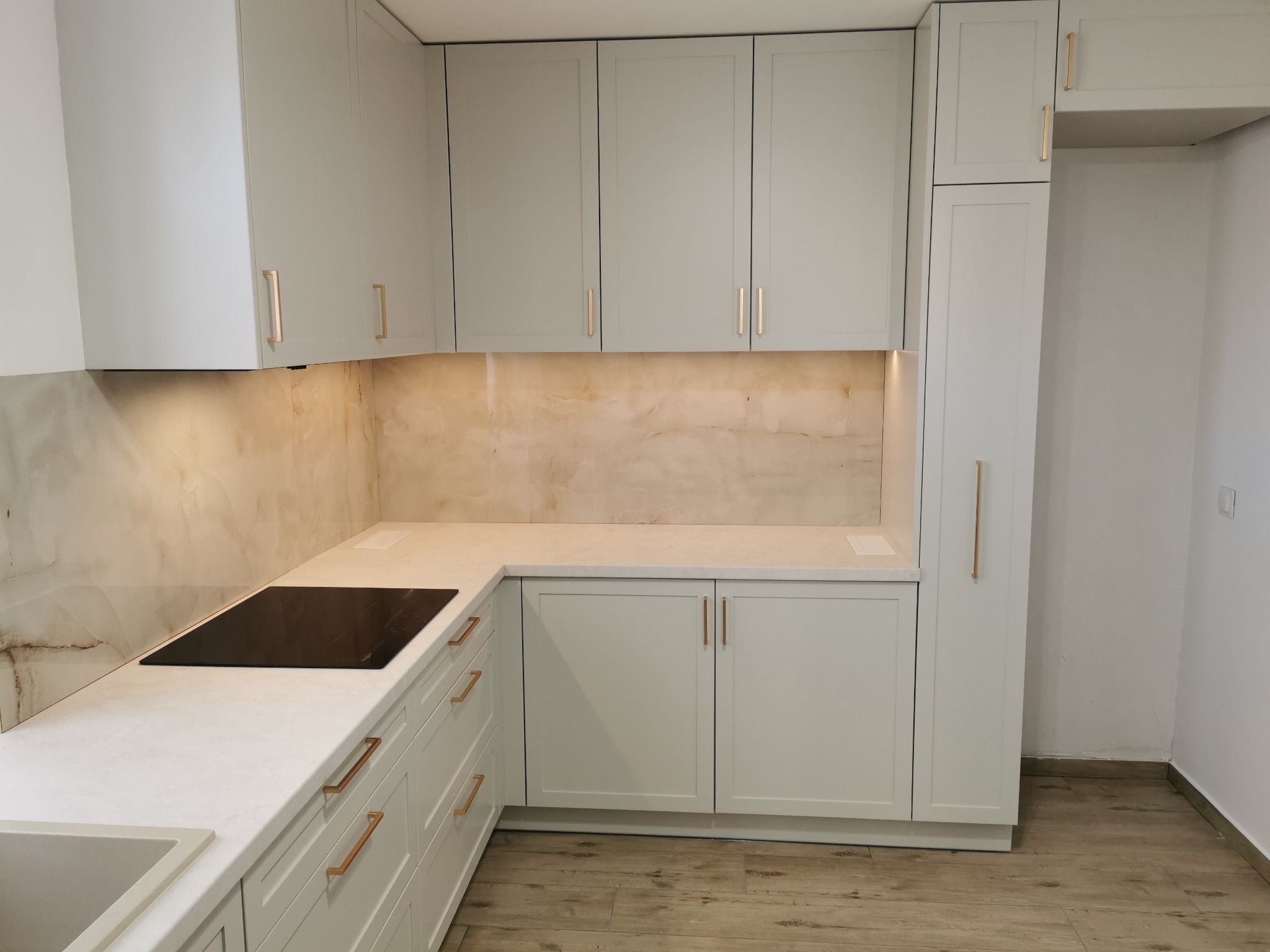 Jasnoszara zabudowa kuchenna z marmurowym blatem i złotymi uchwytami, płyta indukcyjna wkomponowana w blat, podświetlana ściana nad blatem.