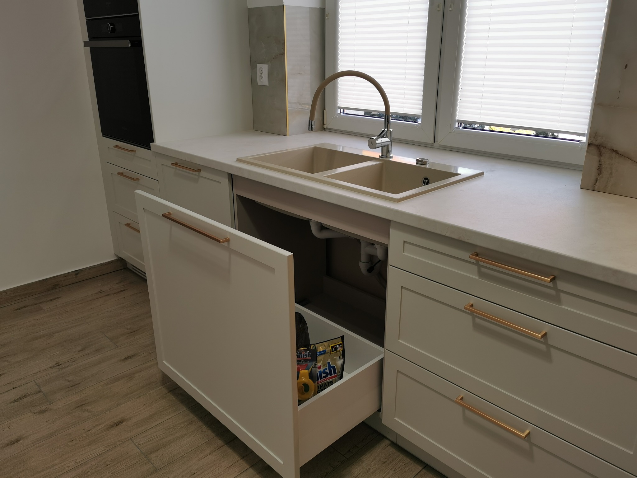 Jasna zabudowa kuchenna w Suwałkach z otwartą szufladą pod zlewem, w której widoczne są środki czystości. Uwagę zwracają złote uchwyty i beżowy zlew z baterią.