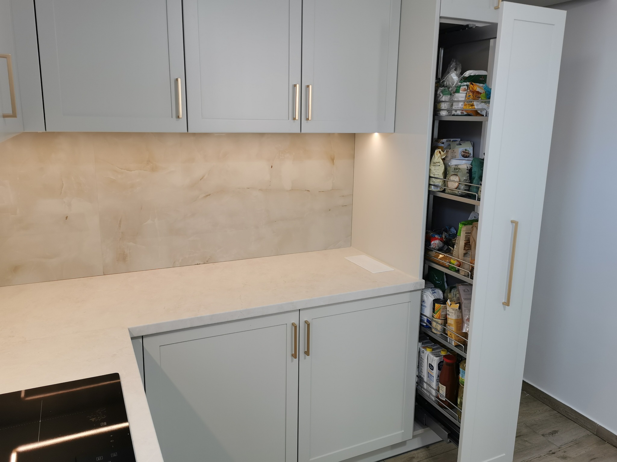 Jasnoszara zabudowa kuchenna z blatem imitującym marmur, wysuwana szafka cargo z zapasami żywności.
