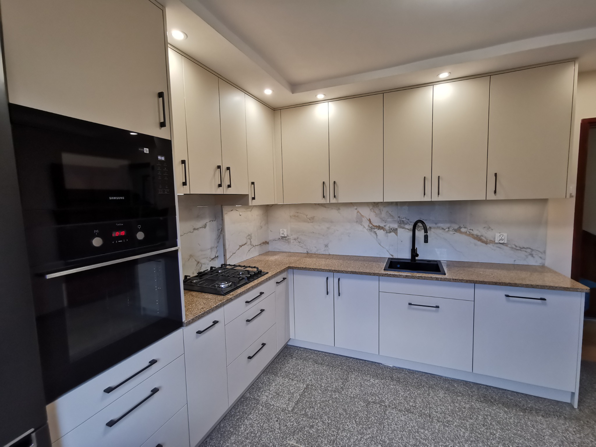 Jasna kuchnia na wymiar z czarnymi detalami: piekarnik Samsung, armatura, uchwyty. Blat w kolorze piaskowym, marmurowe wykończenie ściany. Nowoczesny design.