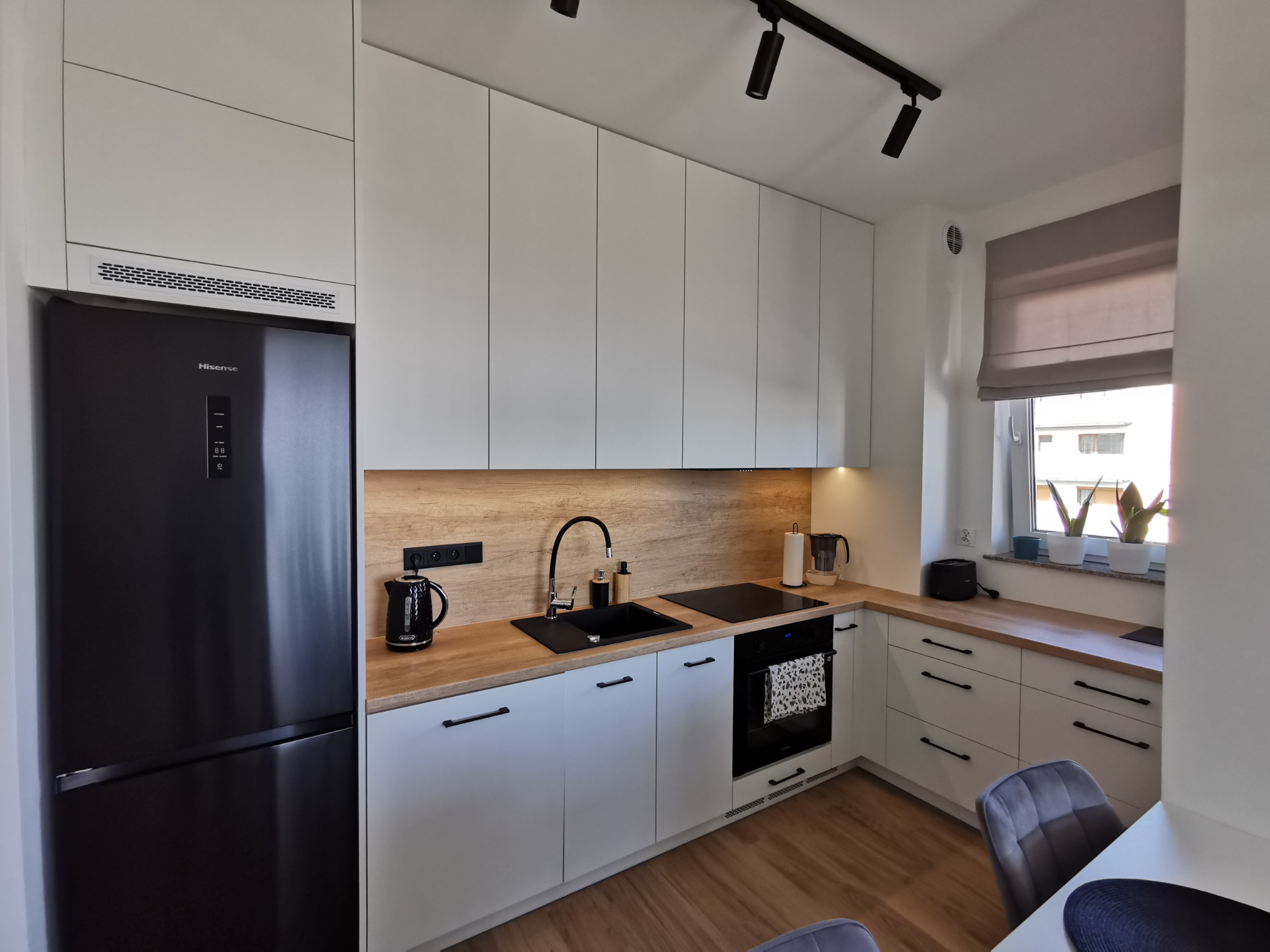 Nowoczesna kuchnia: białe szafki z czarnymi uchwytami, drewniany blat, czarny zlew i lodówka. Nad blatem lampy szynowe. Całość oświetlona, z roślinami w oknie.