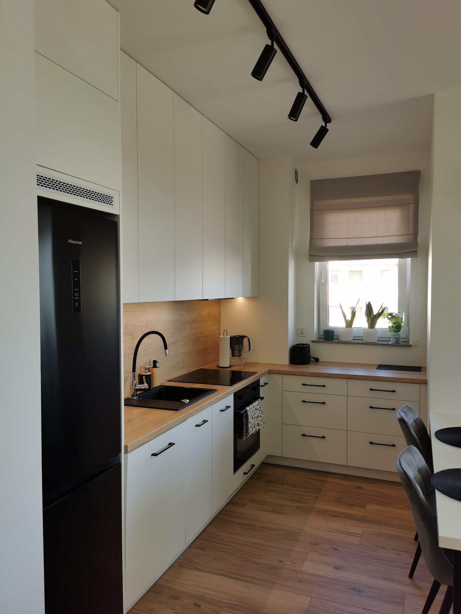 Nowoczesna kuchnia z białymi frontami i drewnianym blatem. Czarny zlew i lodówka. Oświetlenie punktowe. Rośliny na parapecie. Minimalistyczny design.