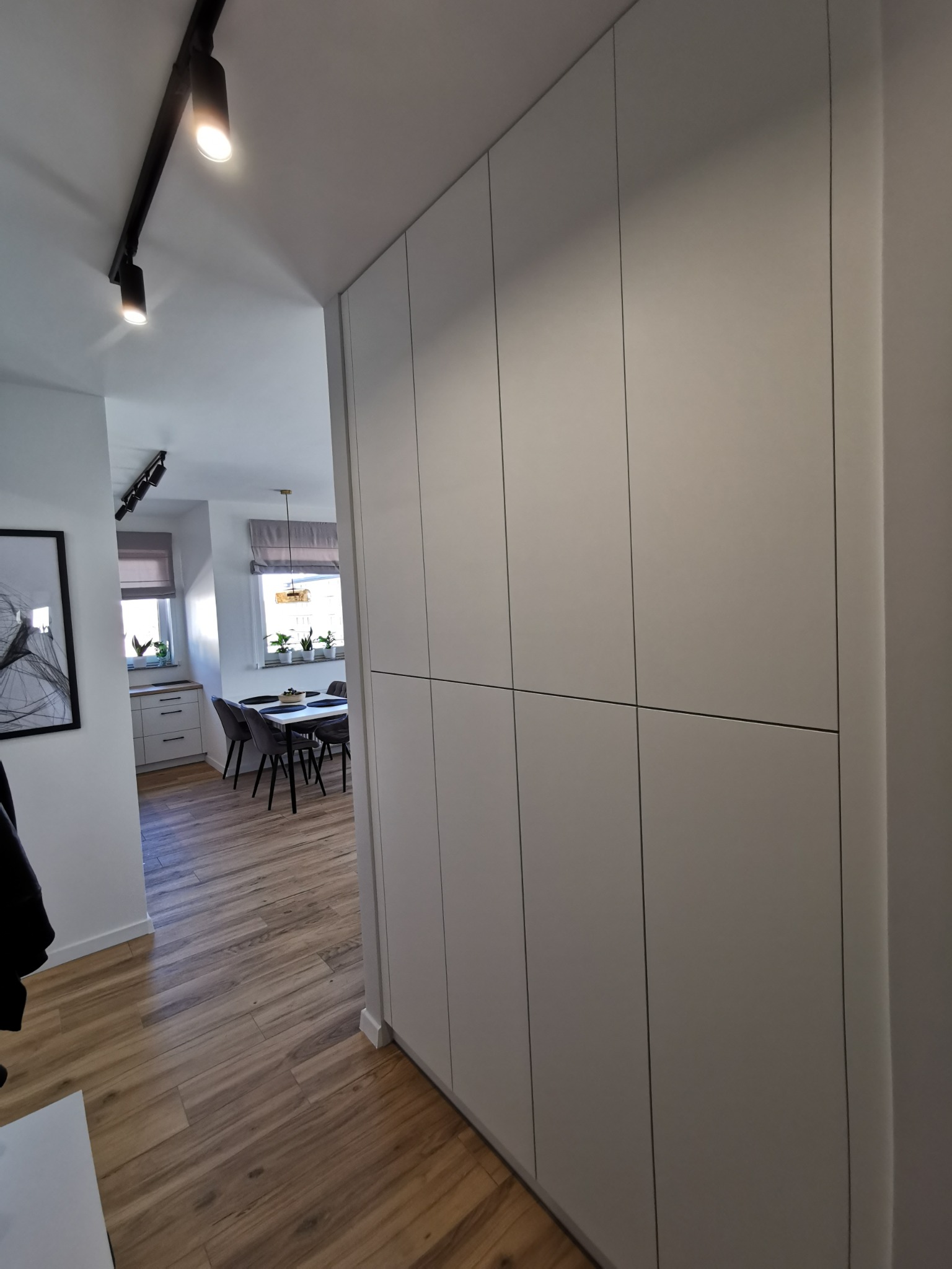 Wysoka, zabudowana szafa wnękowa w przedpokoju, z widokiem na jadalnię. Minimalistyczny design, jasne fronty, oświetlenie szynowe na suficie. Parkiet ułożony w jodełkę.