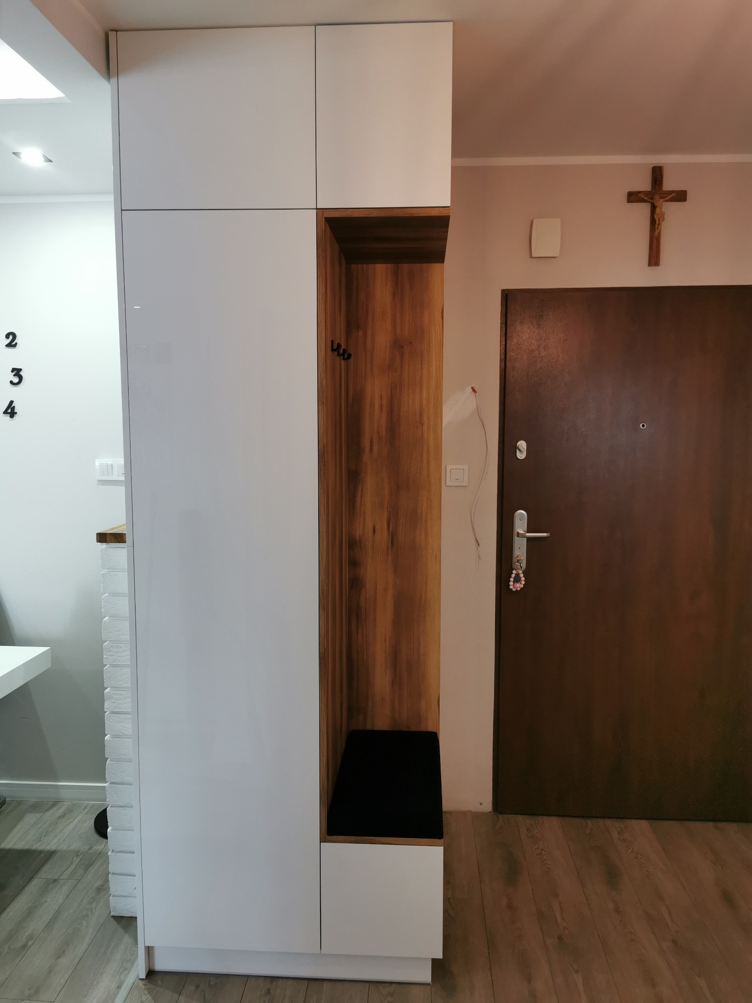 Biała szafa wnękowa z drewnianym siedziskiem i wieszakami w przedpokoju, obok brązowe drzwi wejściowe. Minimalistyczny design, jasne wnętrze.
