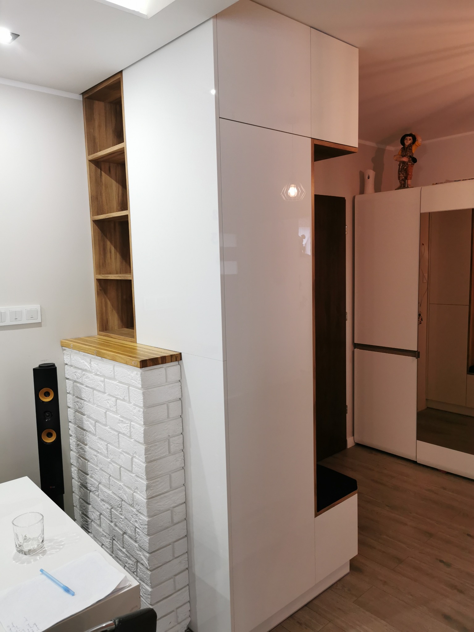 Biała szafa wnękowa z drewnianymi półkami i siedziskiem, połączona z elementem dekoracyjnym z białej cegły, tworząca spójną zabudowę w nowoczesnym wnętrzu.