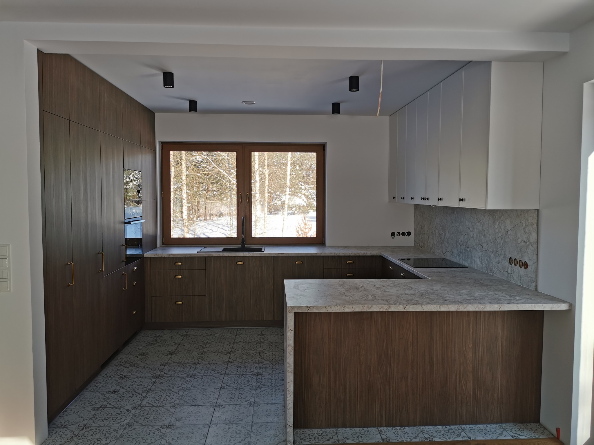 Nowoczesna kuchnia z drewnianymi frontami i marmurowym blatem, widok na zimowy krajobraz przez okno. Uwagę przykuwają złote uchwyty i subtelne oświetlenie.