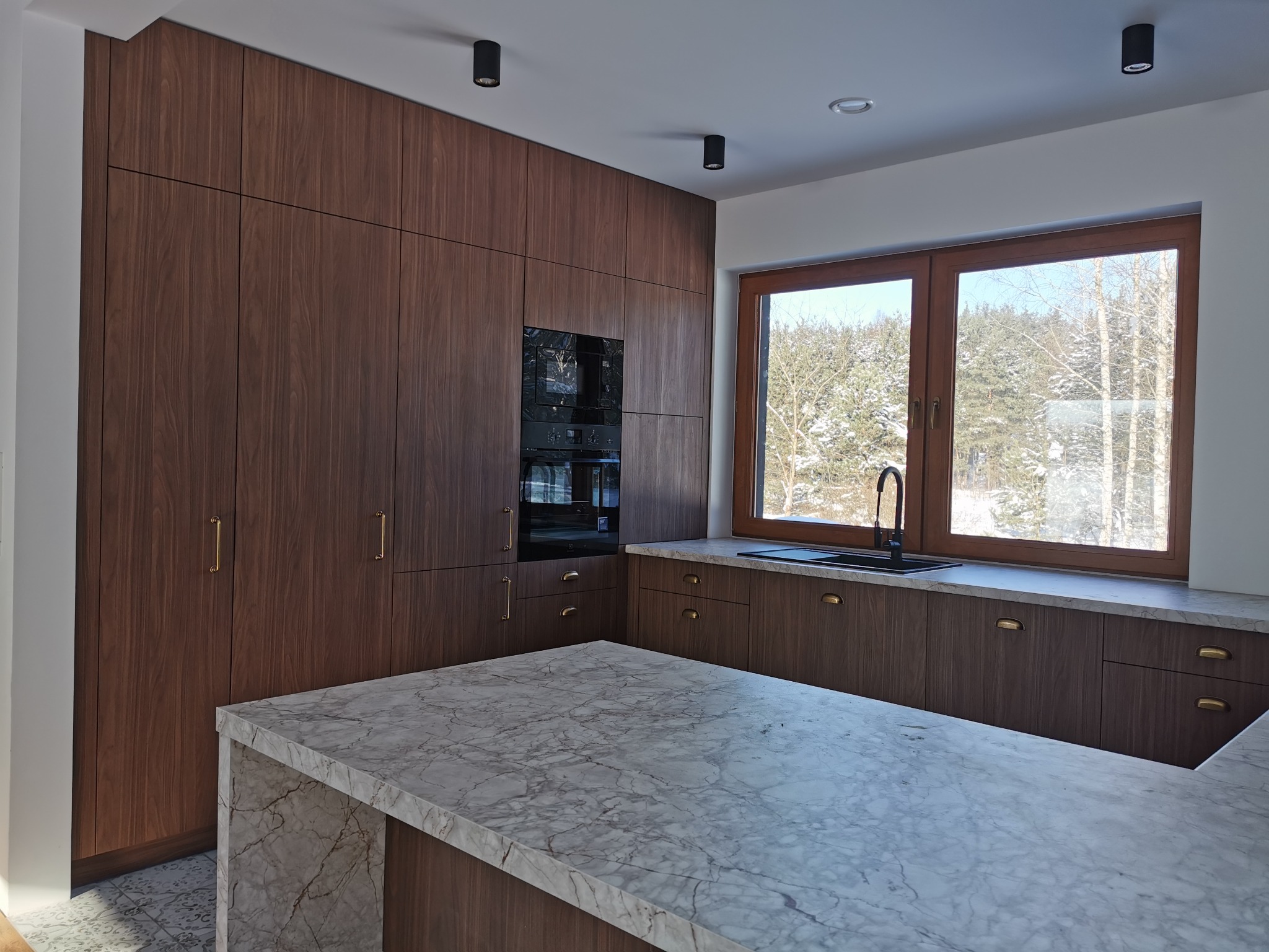Nowoczesna kuchnia z ciemnego drewna z marmurowym blatem wyspowym i złotymi uchwytami, oświetlona punktowo, z widokiem na zimowy krajobraz za oknem.