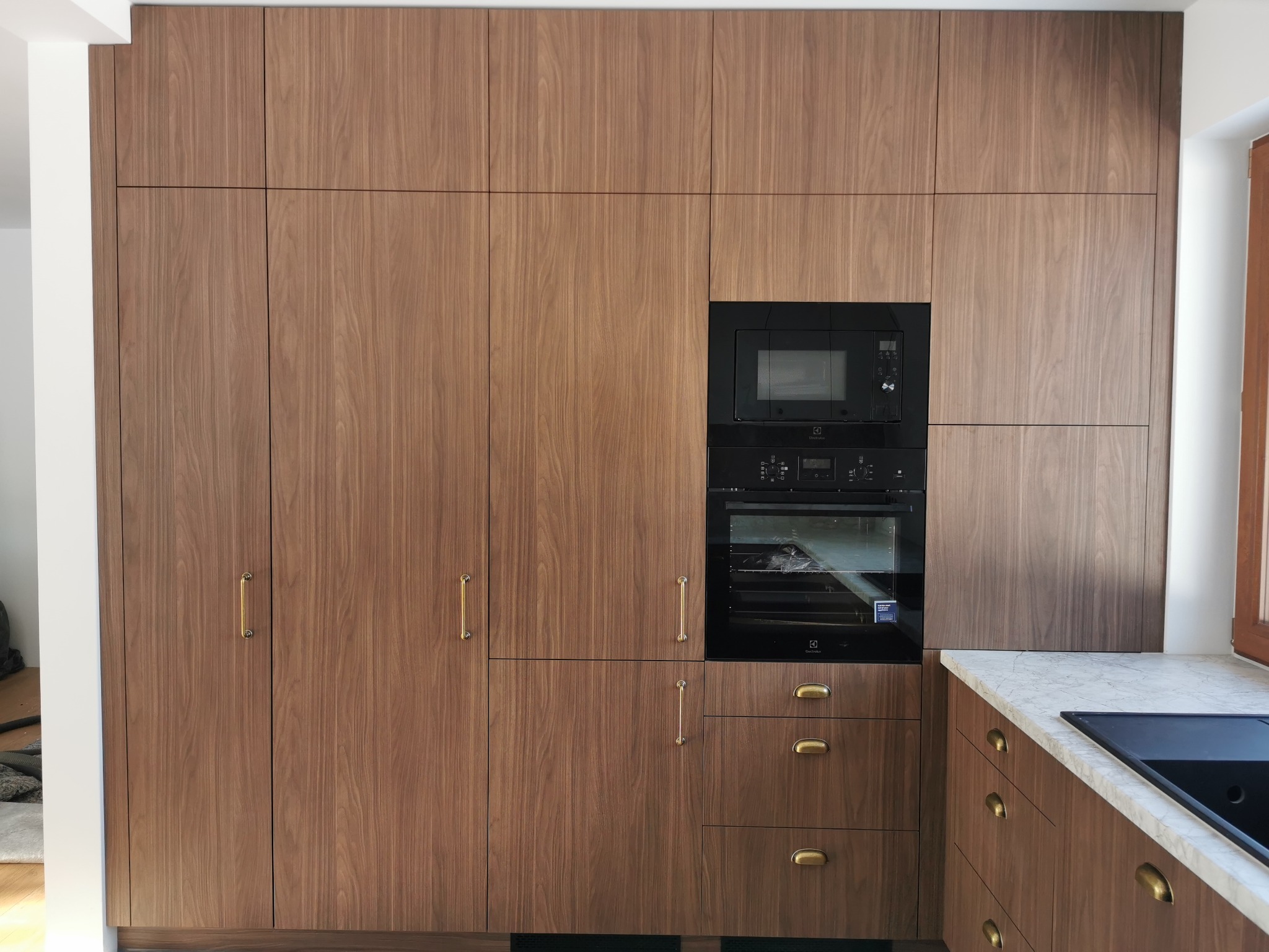 Nowoczesna zabudowa kuchenna z forniru orzechowego, zintegrowana z piekarnikiem i mikrofalówką Electrolux. Minimalistyczne złote uchwyty, marmurowy blat.