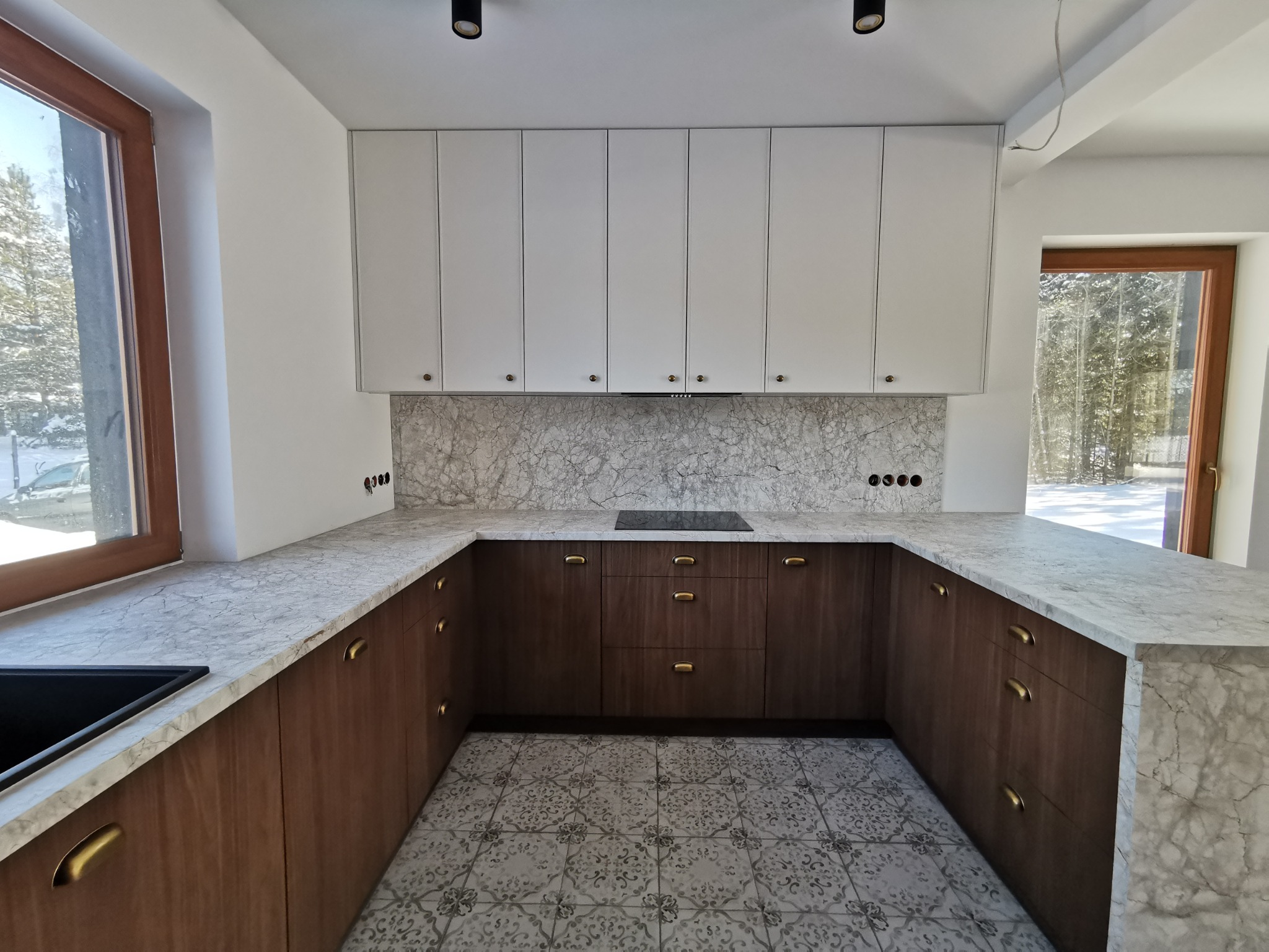 Nowoczesna kuchnia w kształcie litery U z białymi szafkami górnymi i drewnianymi dolnymi, marmurowym blatem i wzorzystą podłogą. Widok na okno z zimowym krajobrazem.