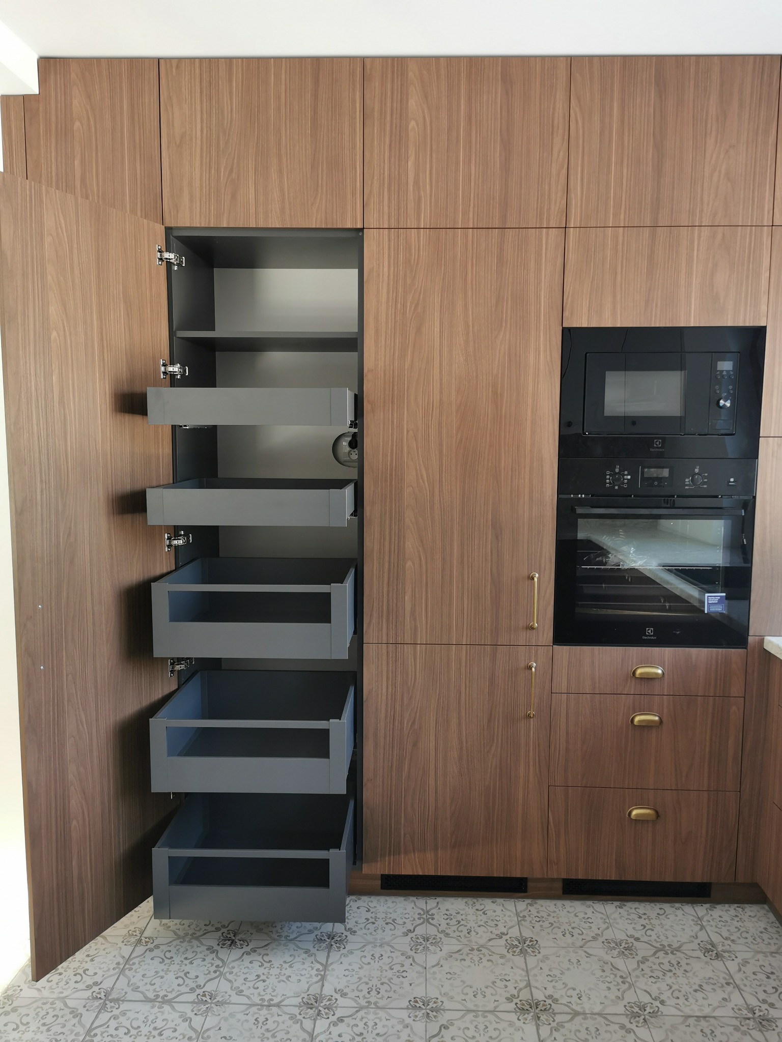 Nowoczesna zabudowa kuchenna z forniru orzechowego, z otwartą szafką cargo i wbudowanym sprzętem AGD. Uwagę przykuwają złote uchwyty i wzorzysta podłoga.