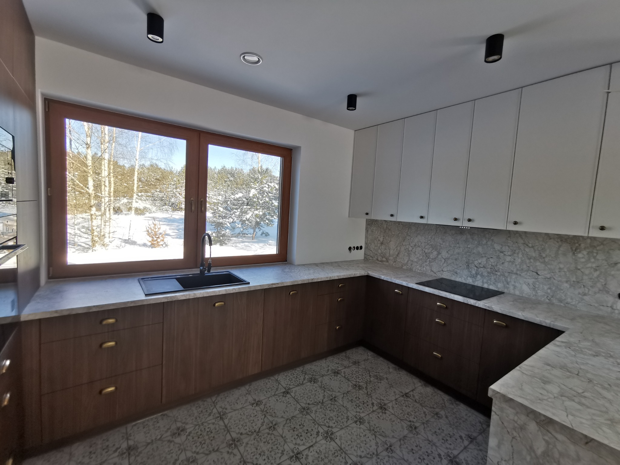 Nowoczesna kuchnia z drewnianymi szafkami i marmurowym blatem, widok na zimowy krajobraz przez okno. Uwagę przyciągają złote uchwyty i wzorzysta podłoga.