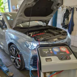 Szary samochód Audi z otwartą maską, zabezpieczony folią ochronną, podłączony do maszyny do serwisowania klimatyzacji R1234yf w warsztacie.