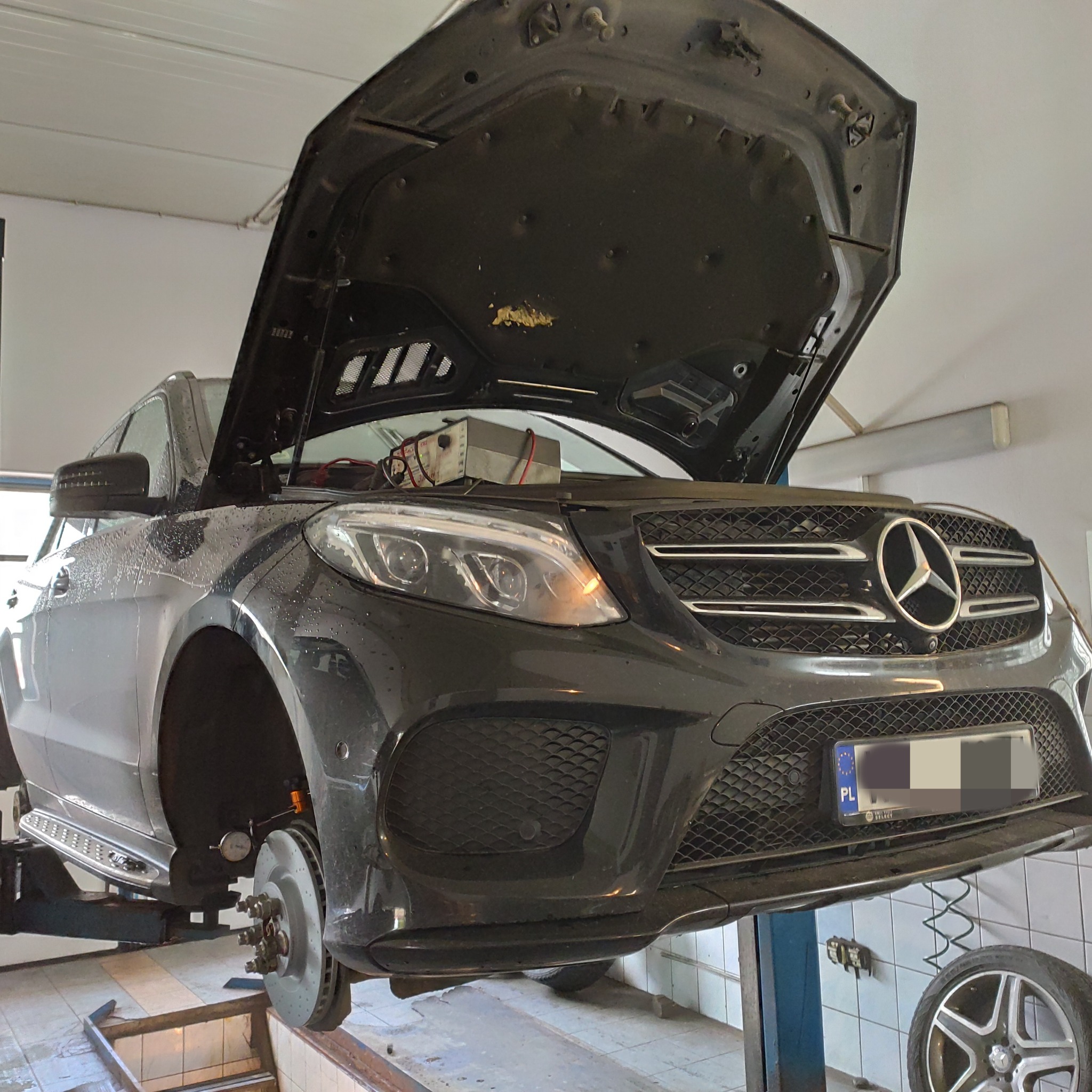 Czarny Mercedes SUV na podnośniku w warsztacie, otwarty silnik, widoczne tarcze hamulcowe i miernik, ładowarka akumulatora podłączona do silnika.
