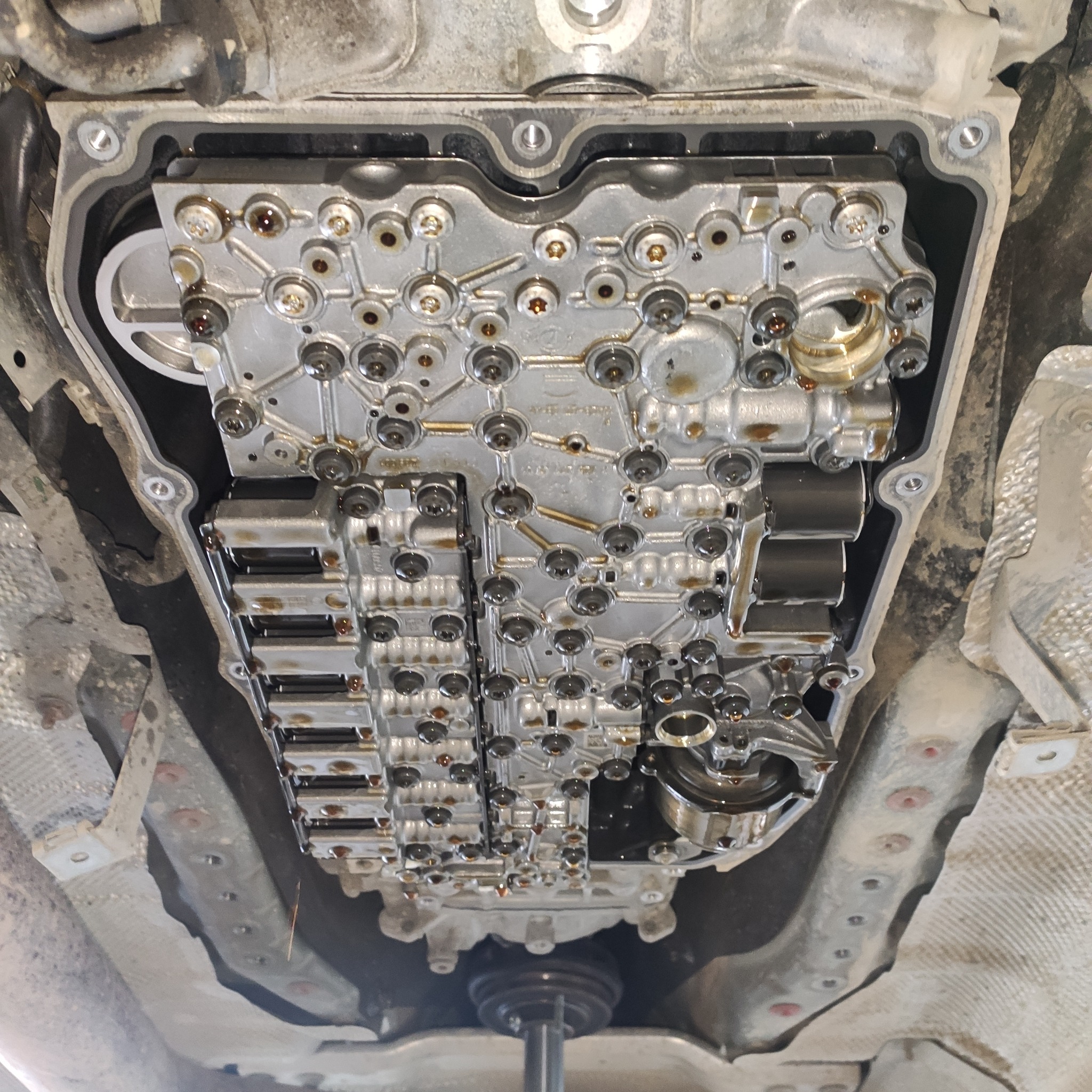 Odkręcona skrzynia biegów automatycznej przekładni z widocznym sterownikiem hydraulicznym, widok od spodu pojazdu.