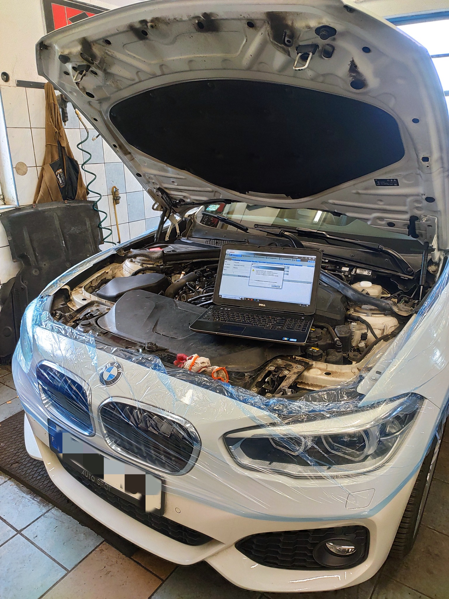 Białe BMW z otwartą maską, zabezpieczone folią, laptop z programem diagnostycznym umieszczony w komorze silnika, widoczne narzędzia i wnętrze warsztatu w tle.