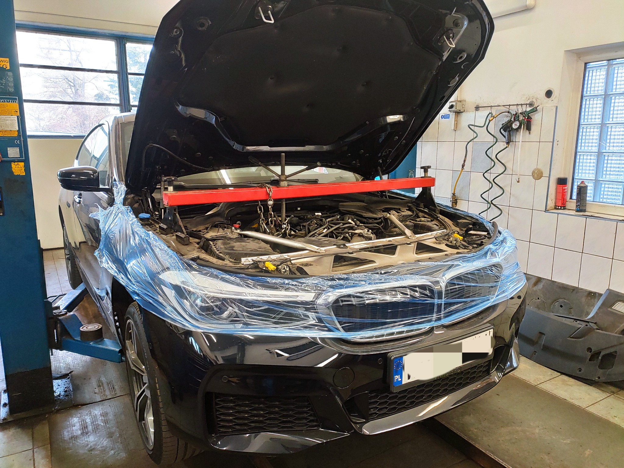 Czarny samochód z otwartą maską, zabezpieczony folią, silnik podtrzymywany czerwoną belką w warsztacie samochodowym.