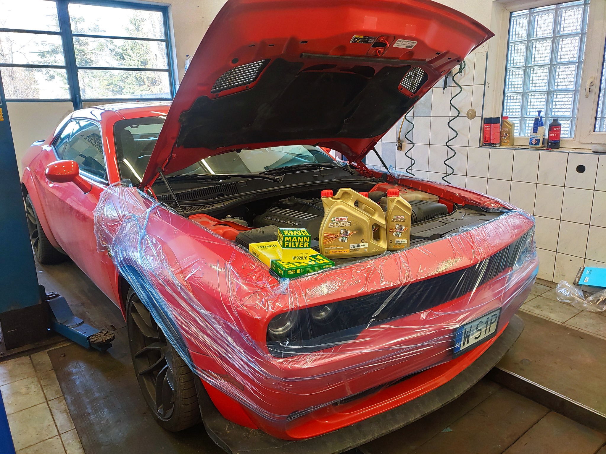 Czerwony samochód sportowy z otwartą maską, zabezpieczony folią ochronną, z filtrami Mann i olejem Castrol Edge na silniku, w tle okno warsztatu.