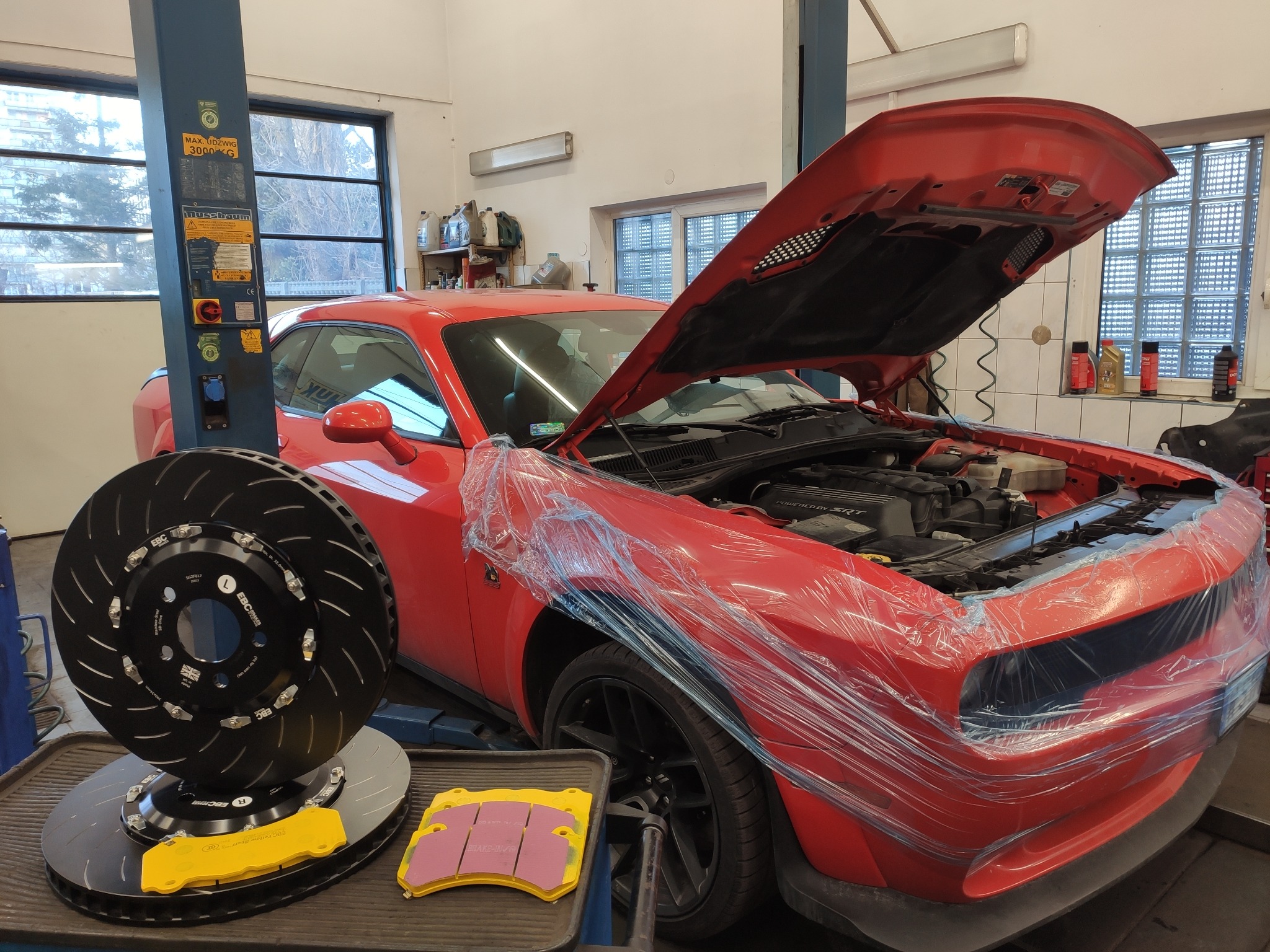 Czerwony samochód sportowy na podnośniku w warsztacie, z otwartą maską i zabezpieczonym przodem folią, obok taca z tarczami hamulcowymi EBC i klockami hamulcowymi