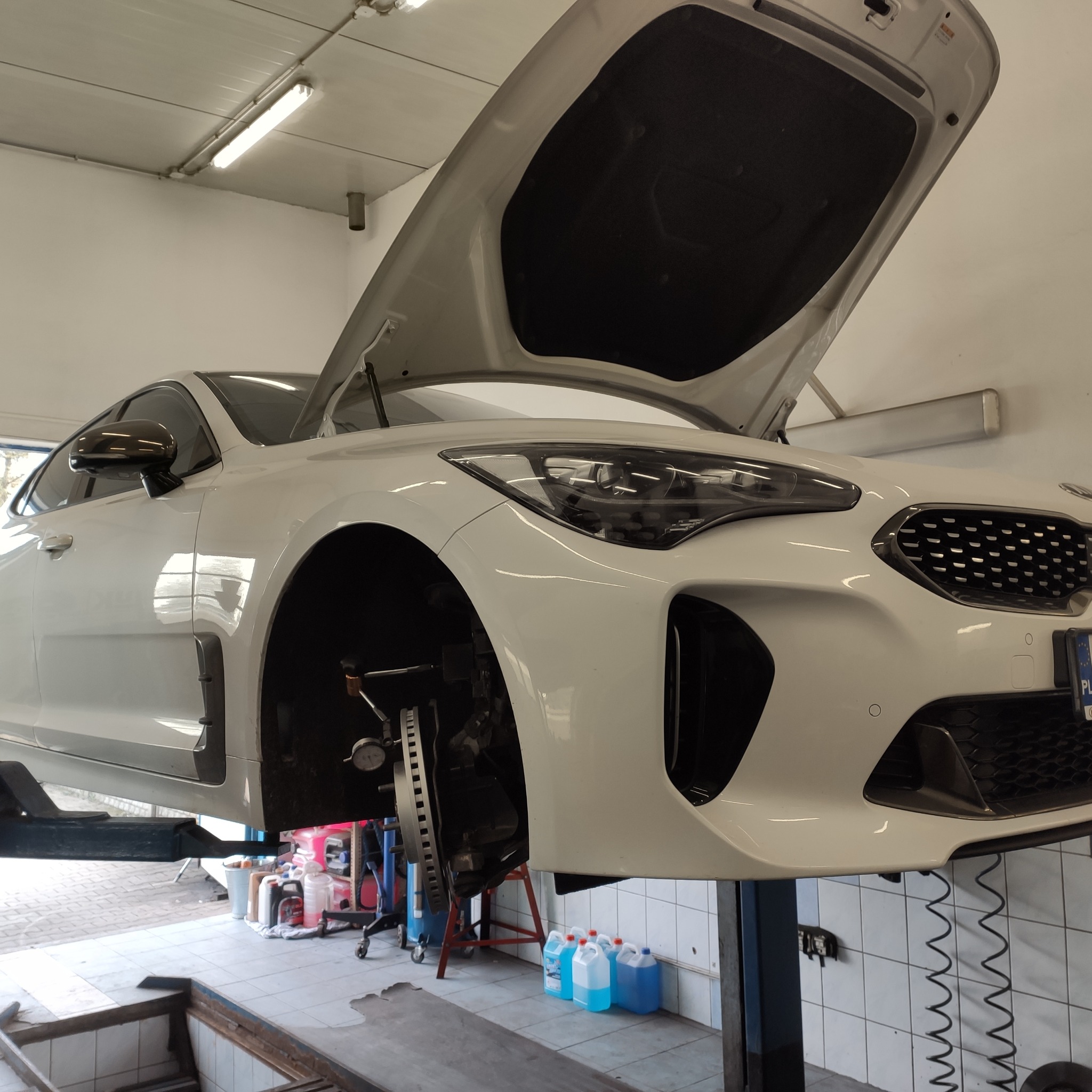 Biały samochód na podnośniku w warsztacie, z otwartą maską i zdjętym kołem, widoczna tarcza hamulcowa i zacisk, w tle plastikowe pojemniki z płynem.