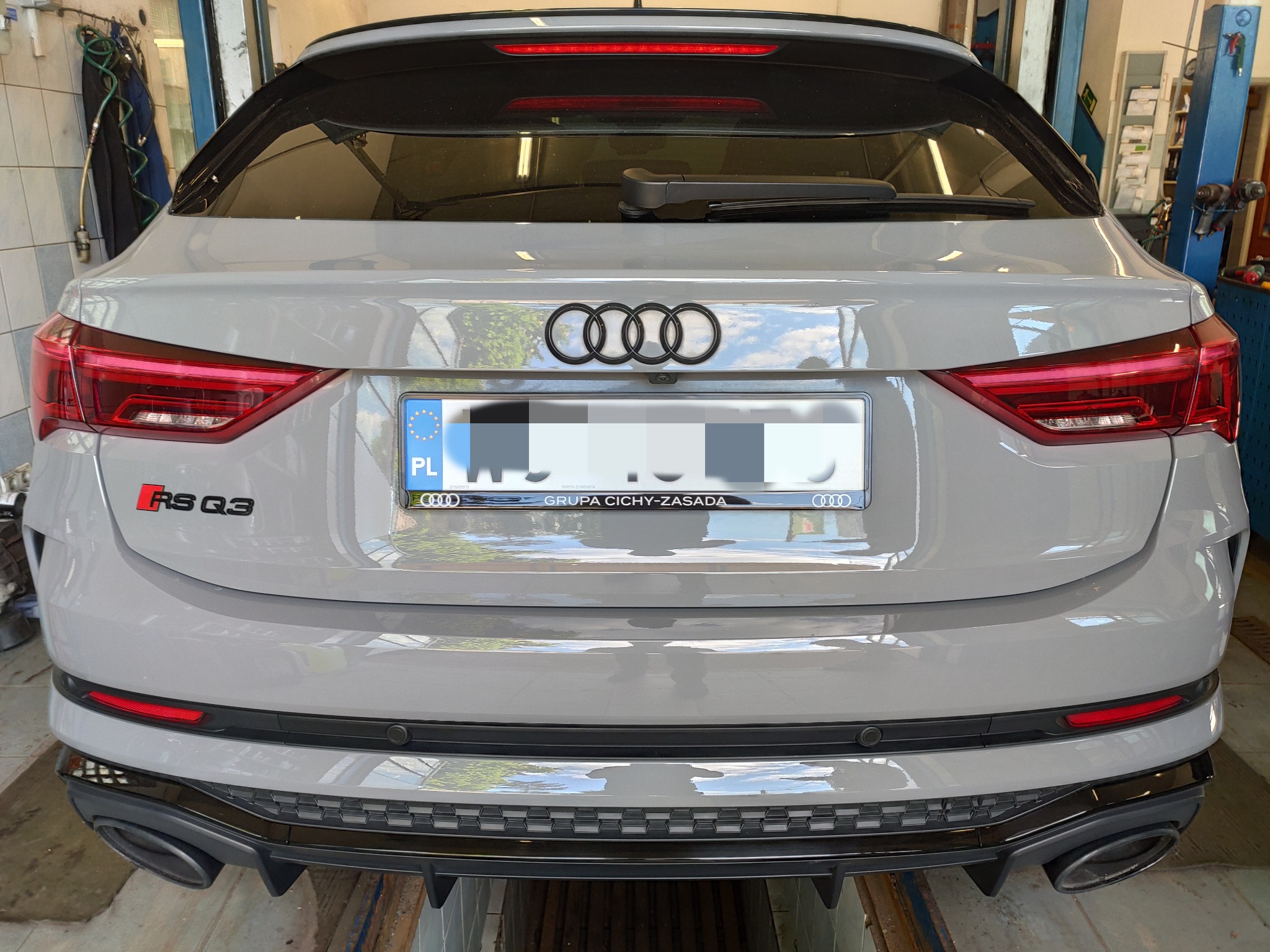 Tył szarego samochodu Audi RS Q3 w warsztacie, widoczne logo Audi, tablica rejestracyjna i dyfuzor z końcówkami wydechu.