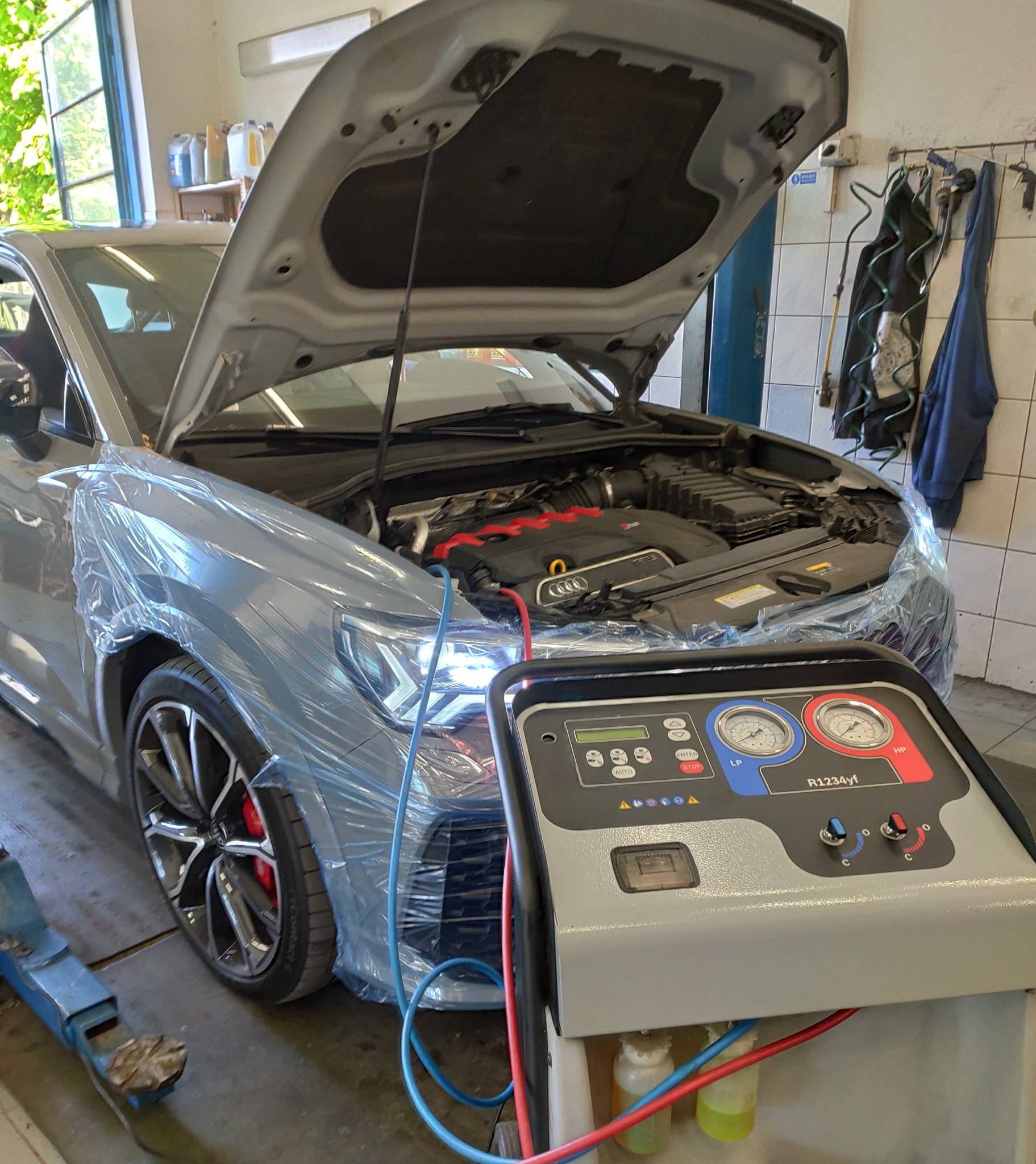 Szary samochód Audi z otwartą maską, zabezpieczony folią ochronną, podłączony do maszyny do serwisowania klimatyzacji R1234yf w warsztacie.