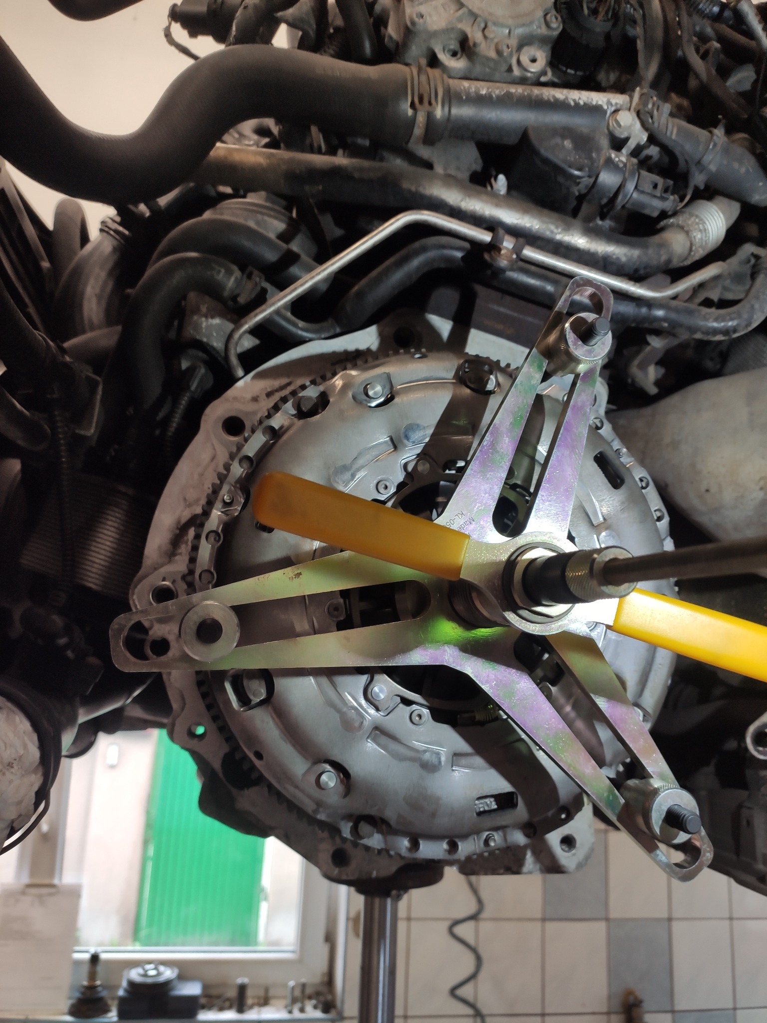 Sprzęgło samochodowe zamontowane do silnika, zabezpieczone specjalistycznym narzędziem z żółtymi elementami, widok od dołu w warsztacie samochodowym z oknem w tle.