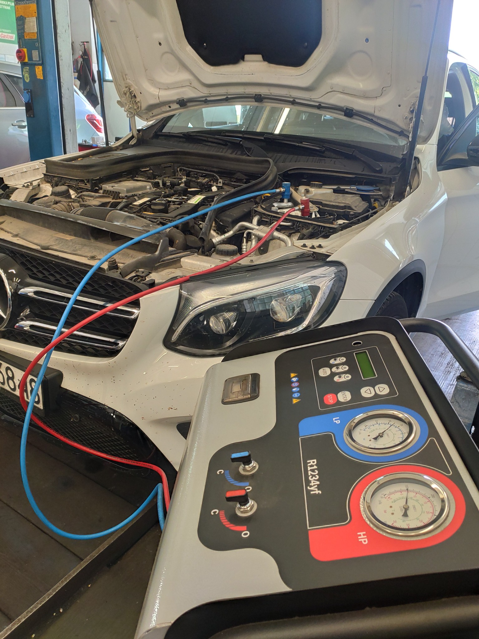 Biały samochód Mercedes z otwartą maską, podłączony do maszyny do napełniania klimatyzacji, widoczne węże i manometry urządzenia R1234yf.