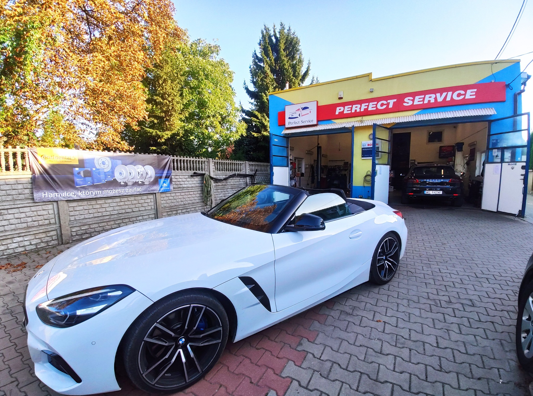 Biały kabriolet BMW Z4 zaparkowany przed warsztatem samochodowym Perfect Service z otwartymi bramami, w tle widoczny podnośnik i inny samochód, na ogrodzeniu baner reklamowy hamulców Continental.